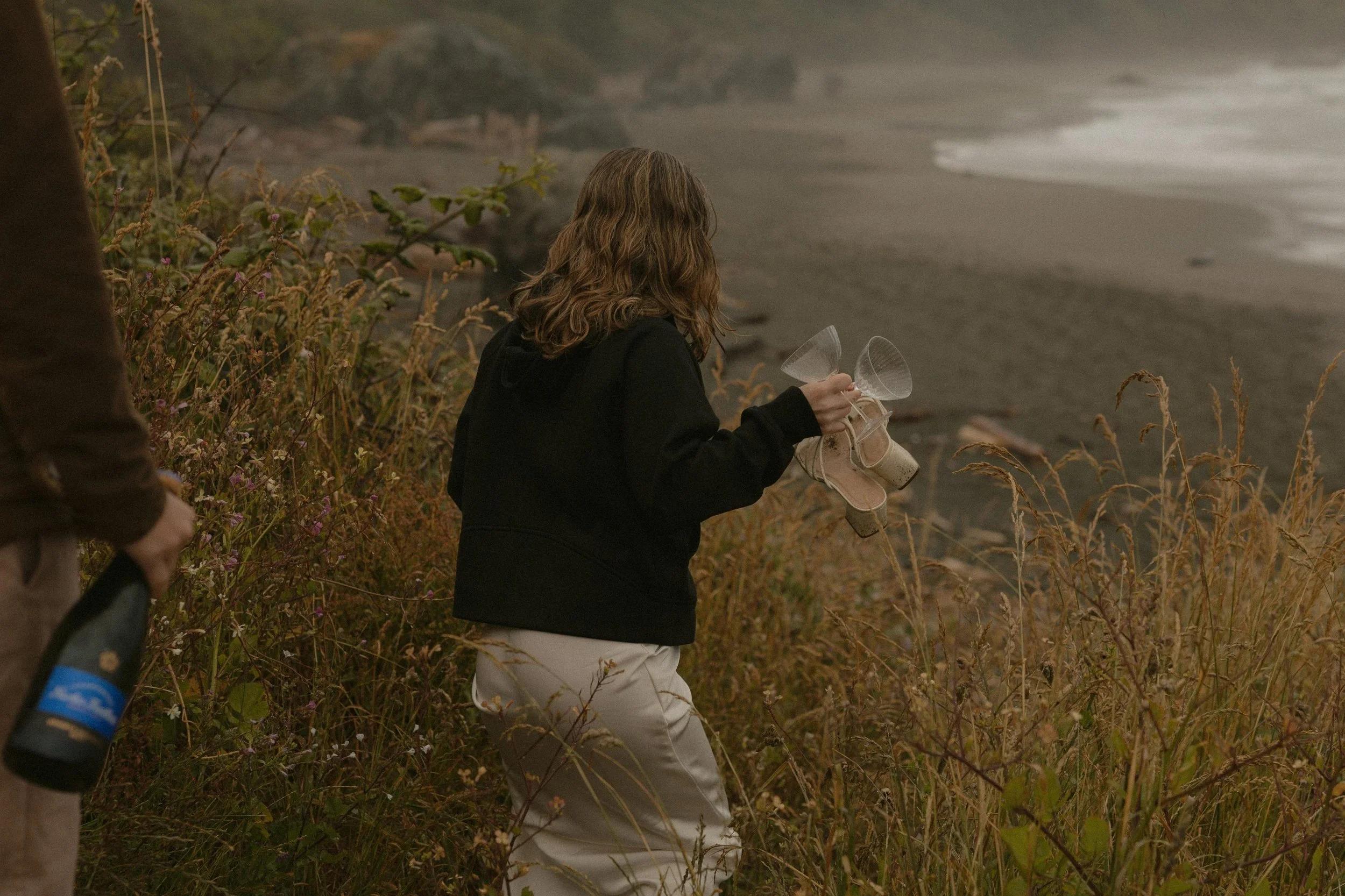 A woman with light brown, wavy hair, wearing a black jacket and white pants, holding a pair of white high-heeled shoes, standing by a beach on a cloudy day.
