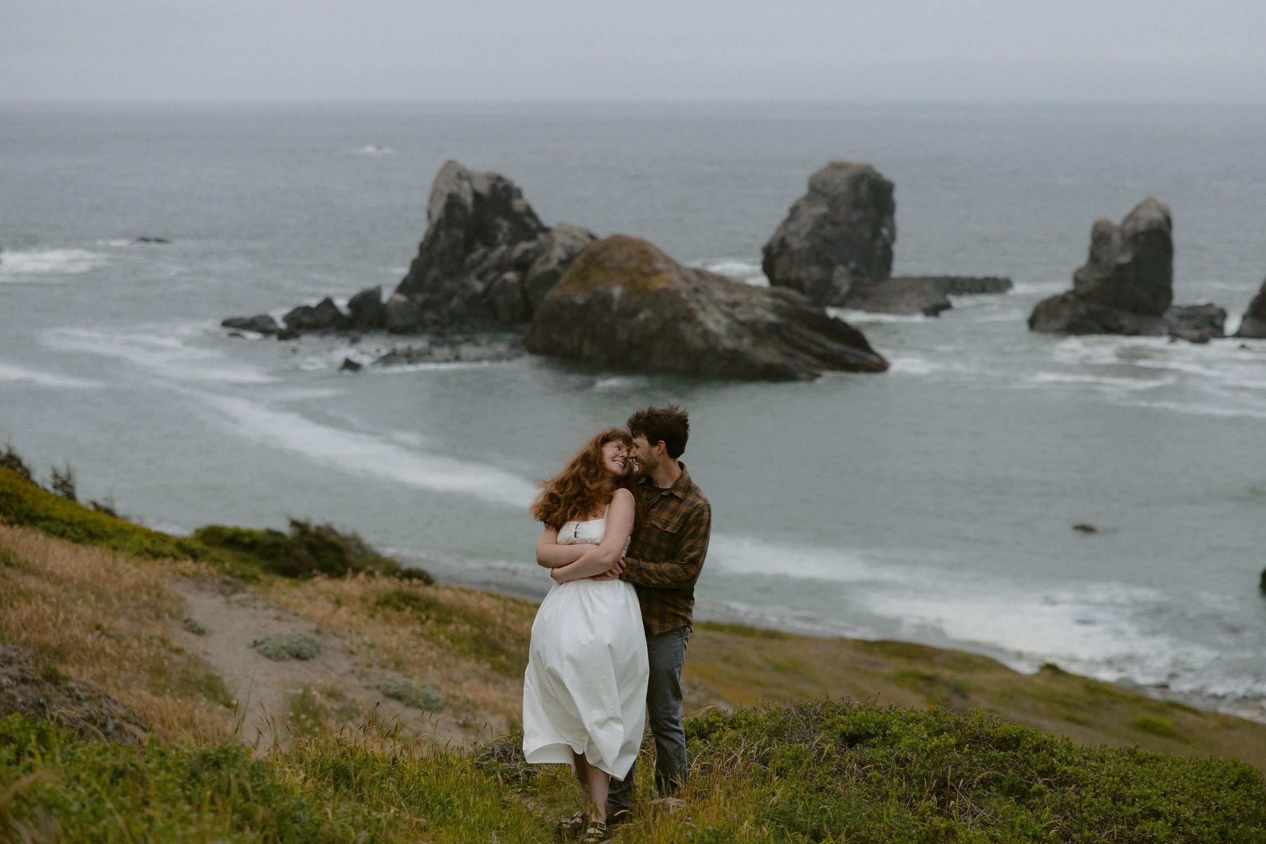 A couple stands close together on a grassy path near the coast, with large rock formations in the ocean behind them, on a cloudy day.