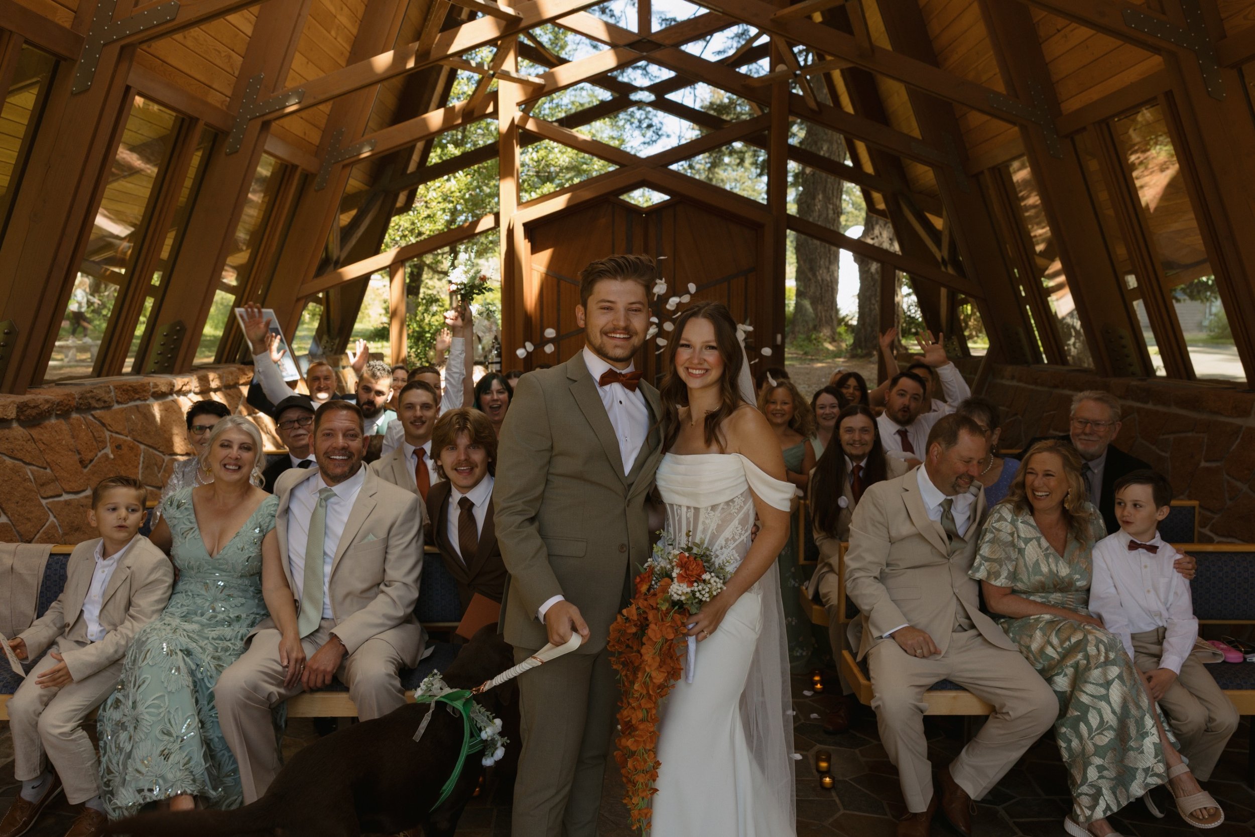A bride and groom standing together at a wedding ceremony, surrounded by seated guests, inside a wooden chapel with glass windows and trees visible outside.