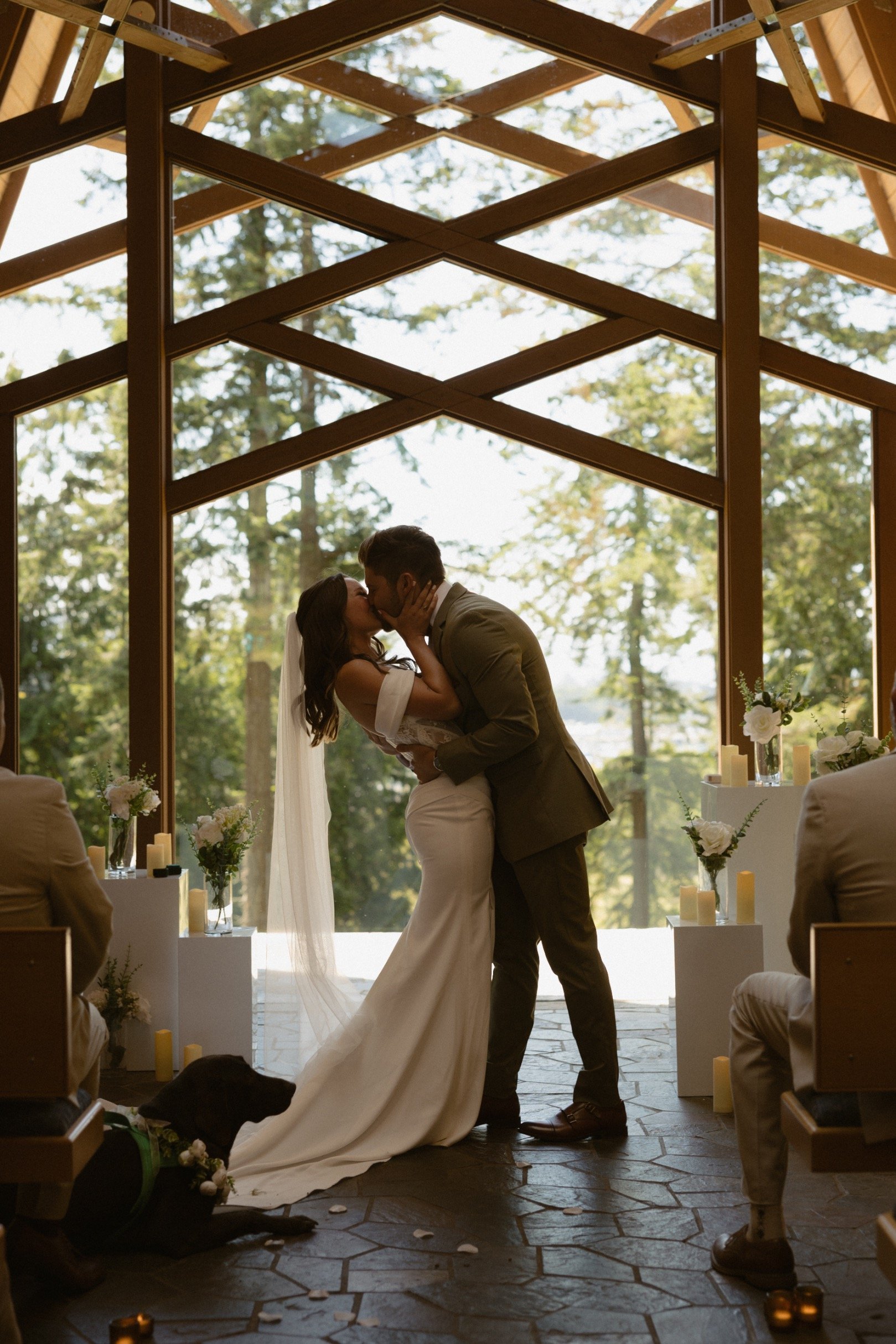 A bride and groom sharing a kiss during their wedding ceremony in a wooden chapel with large glass windows and nature in the background, surrounded by candles and flower arrangements.