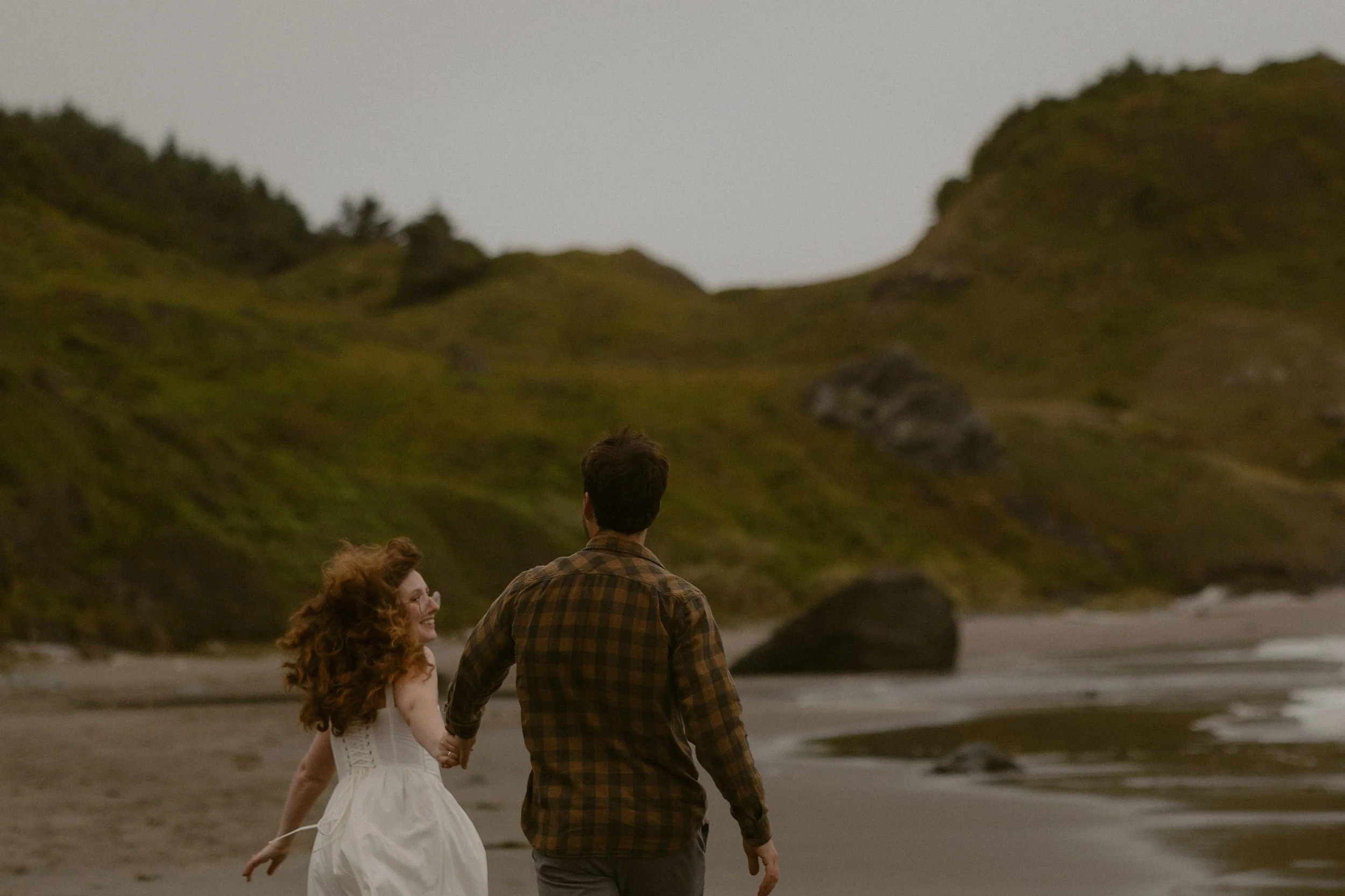 A group of three people, two women and one man, are walking hand in hand along the beach at sunset or dusk, with green hills in the background.