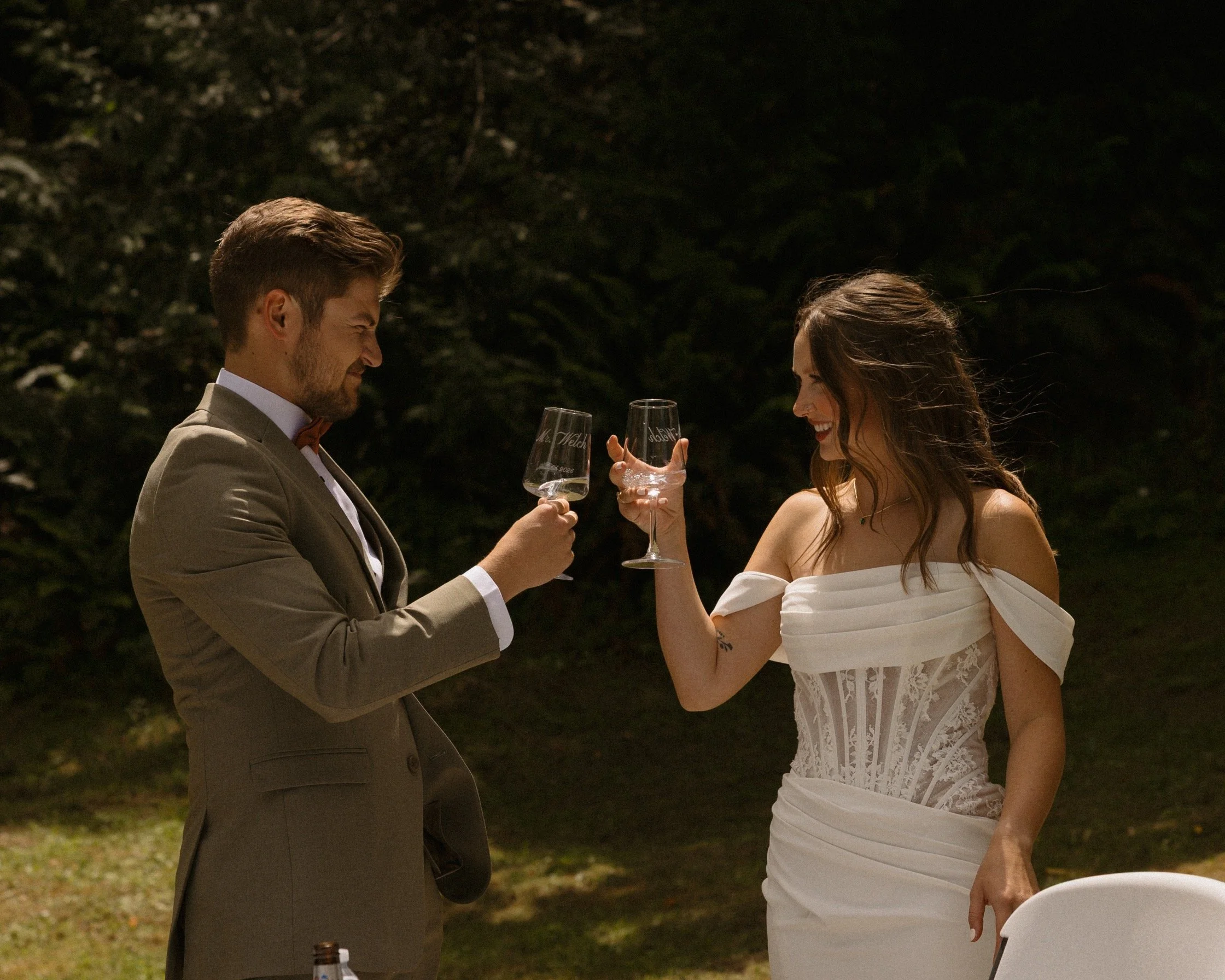 A man and woman dressed in formal attire are smiling and toasting with wine glasses at an outdoor event during nighttime.