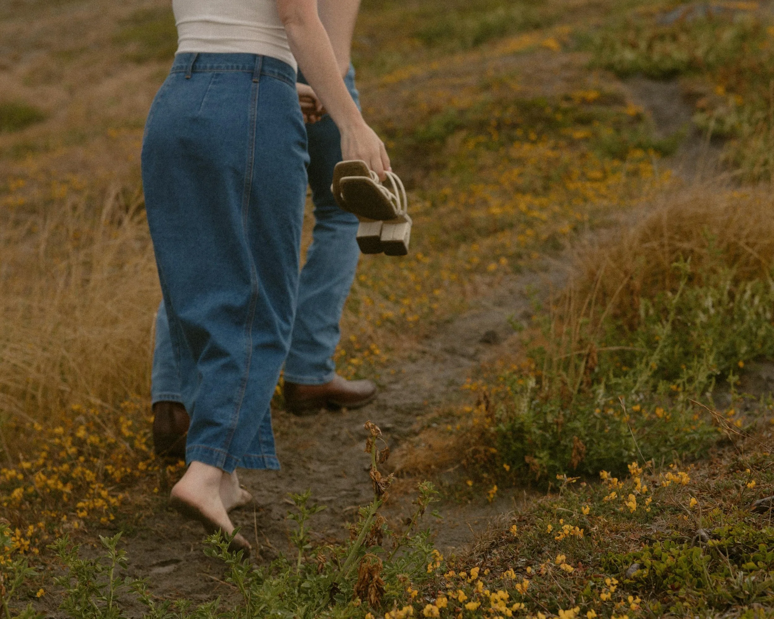 Person standing barefoot on dirt path, holding brown hiking shoes with white laces in hand. Wears blue jeans rolled up at the ankle and a light-colored sleeveless top. Surrounds are grassy and dotted with small yellow flowers, with a wooded area in t