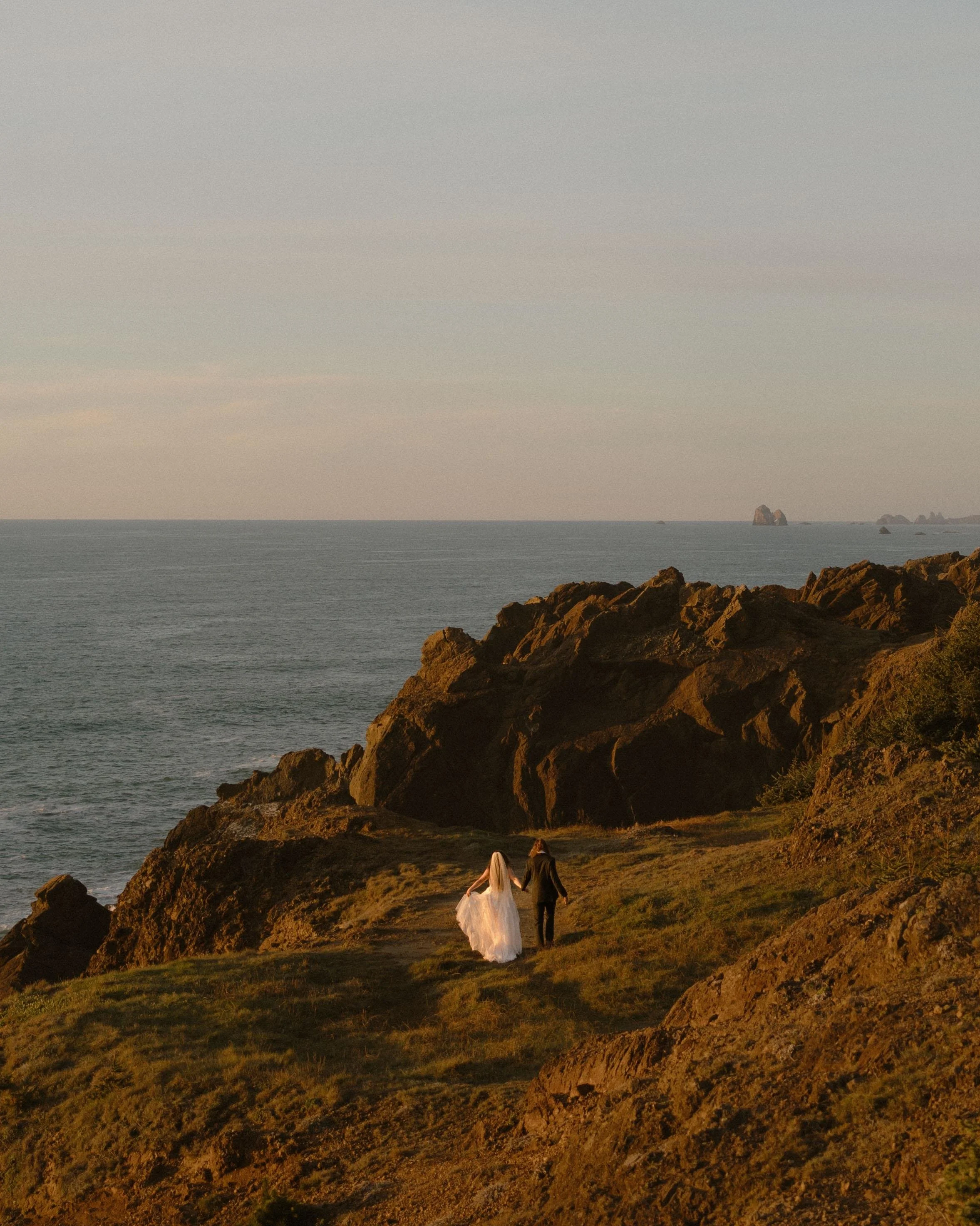 A couple walking hand in hand along a rocky coastline during sunset, with the ocean in the background and a couple in wedding attire.