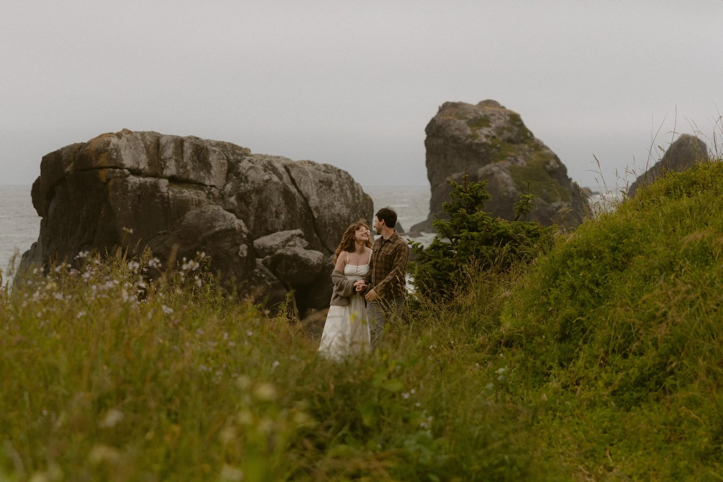 A couple standing close together on a grassy area by the ocean, with large rocks and cliffs in the background, during overcast weather.