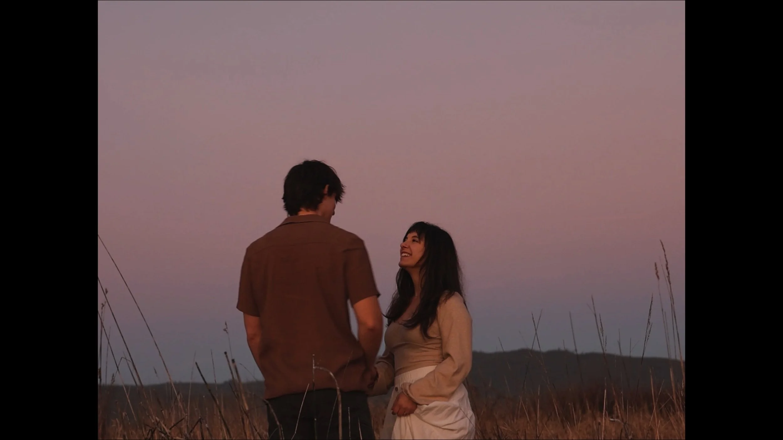 A man and woman standing outdoors in a field at sunset, looking at each other and smiling.