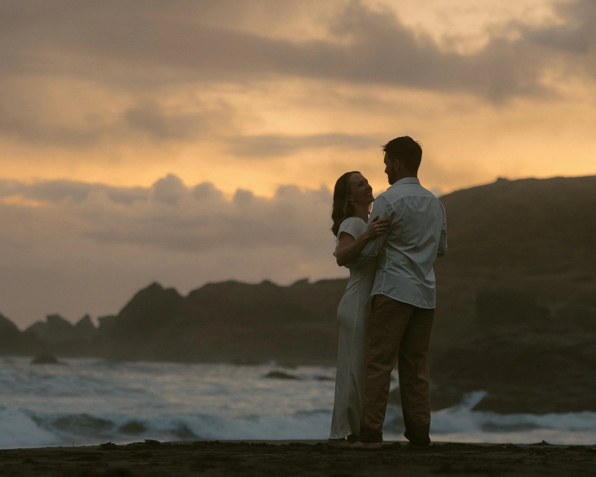 A couple standing close together on a beach at sunset, gazing into each other's eyes with the ocean waves in the background.