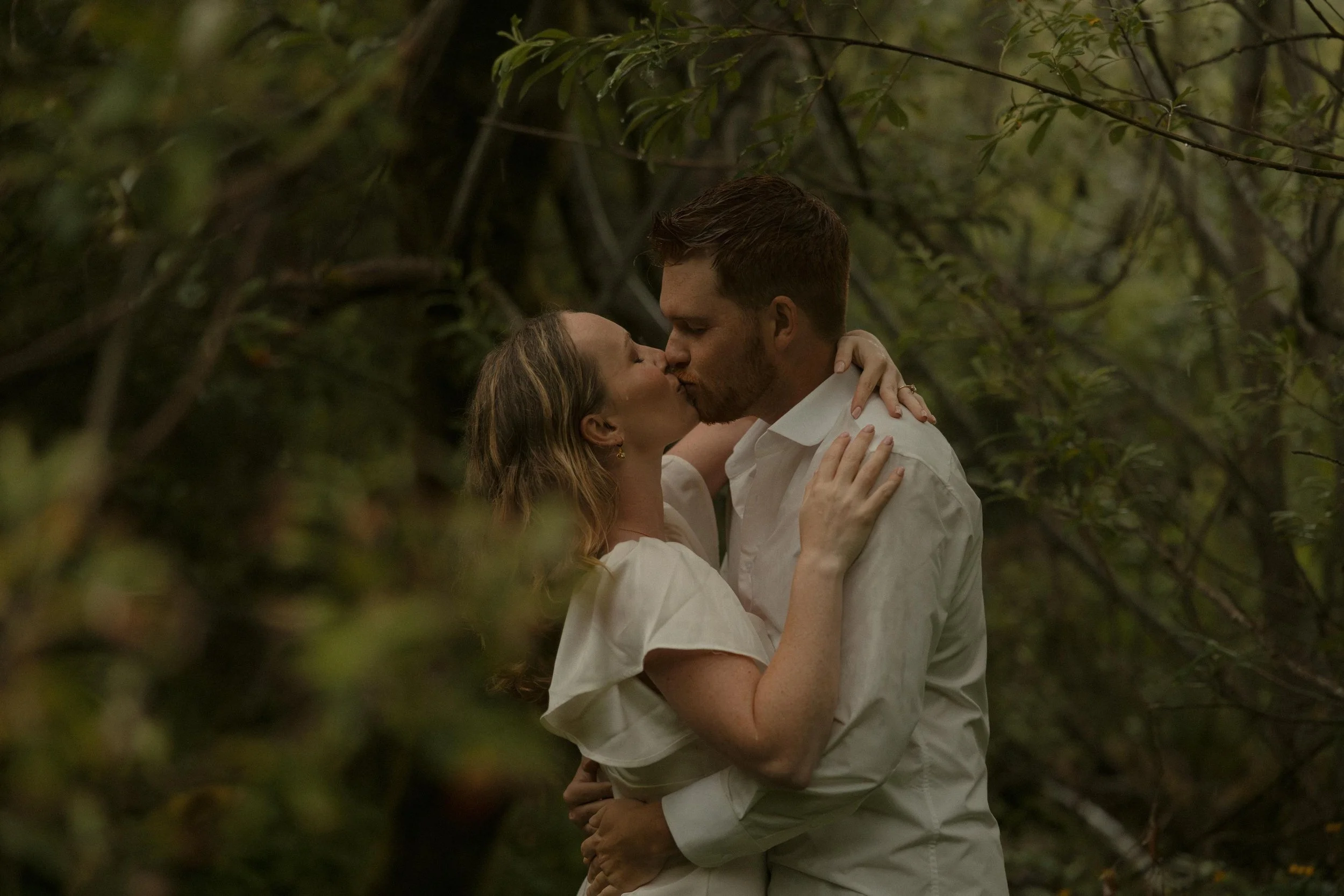 A couple kissing amidst dense green foliage.