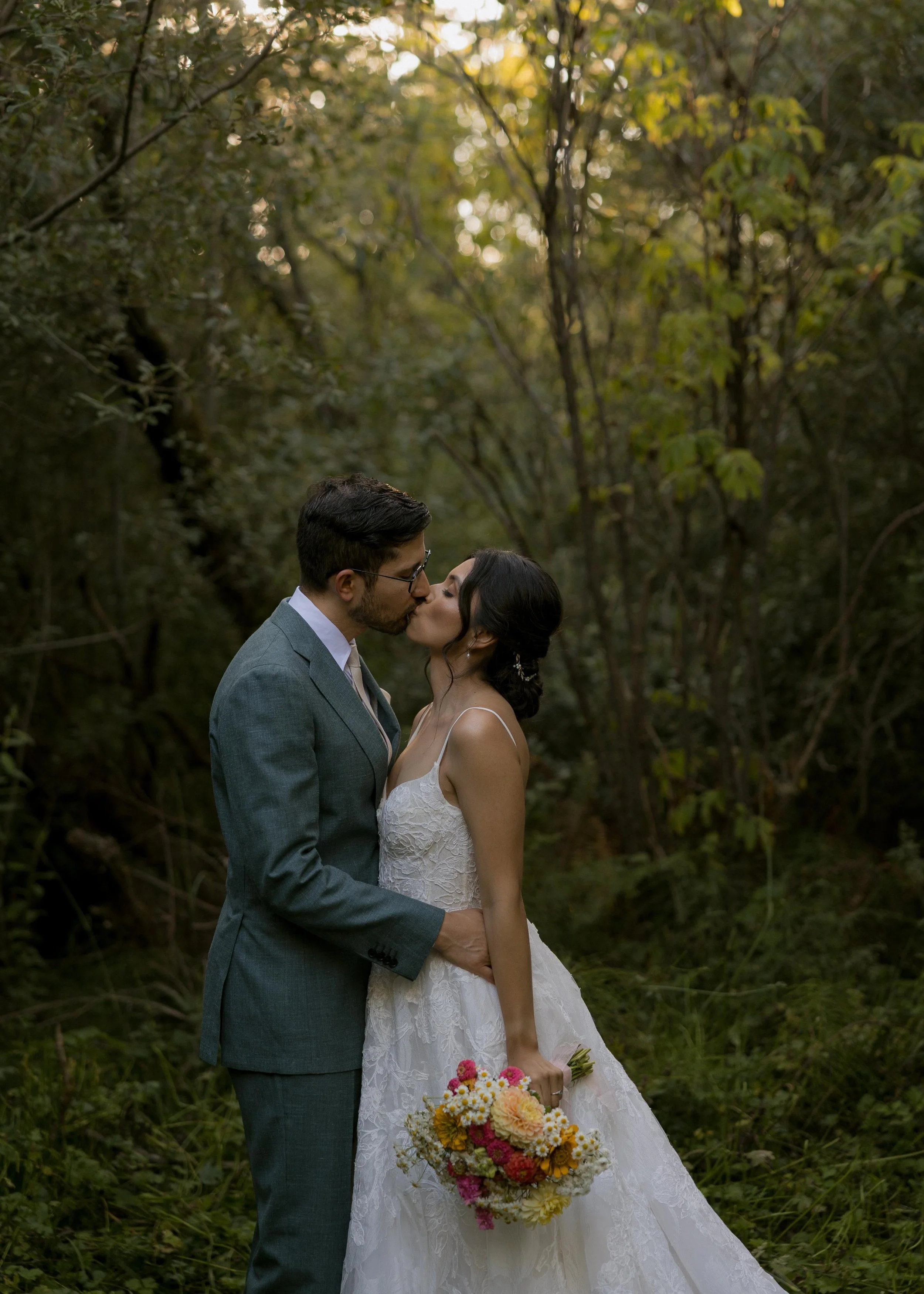 A bride and groom sharing a kiss in a forest, with the bride holding a vibrant bouquet of flowers.
