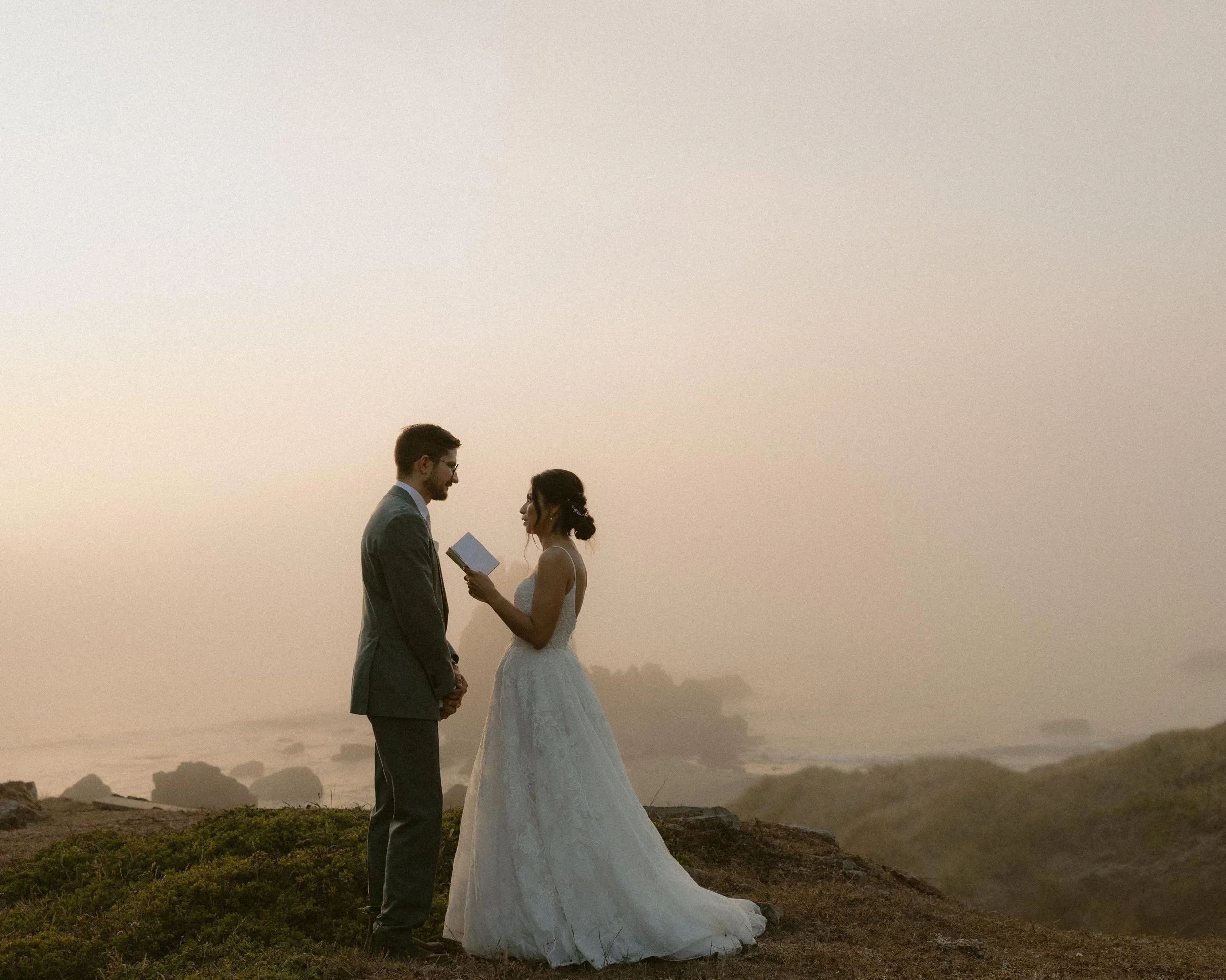 A bride and groom standing face to face outdoors at sunset or sunrise, with barren landscape and rocky terrain in the background.