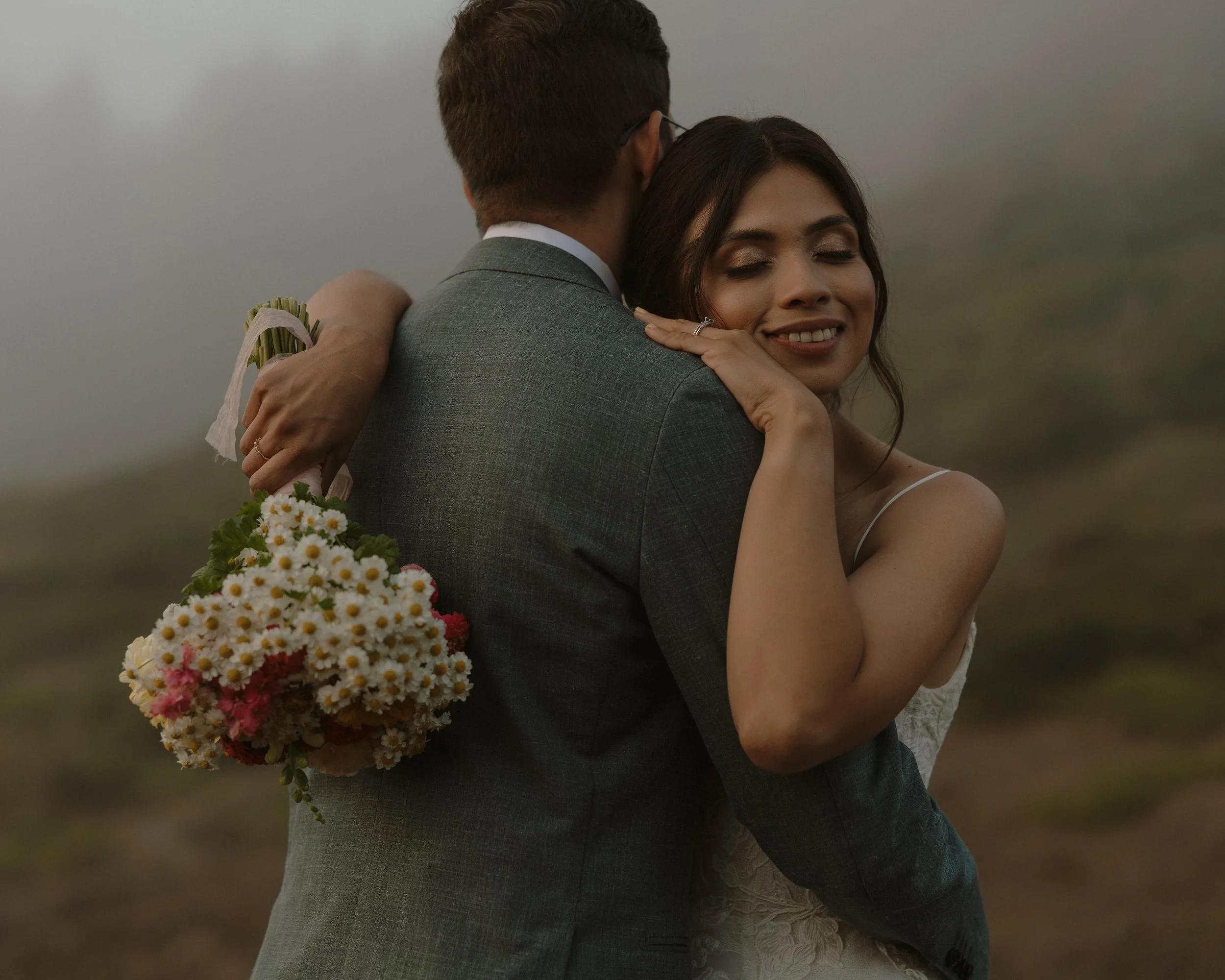 A couple embracing, with the woman smiling and holding a bouquet of flowers, in an outdoor setting.