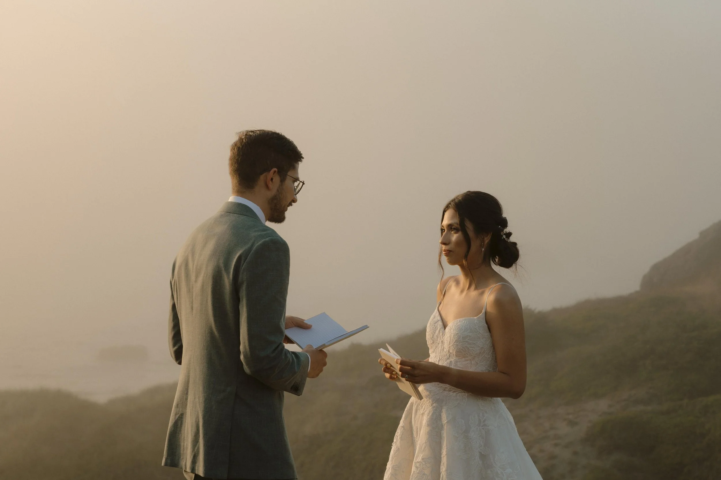 A man and woman dressed in wedding attire are standing outdoors at sunset, exchanging vows and holding papers.