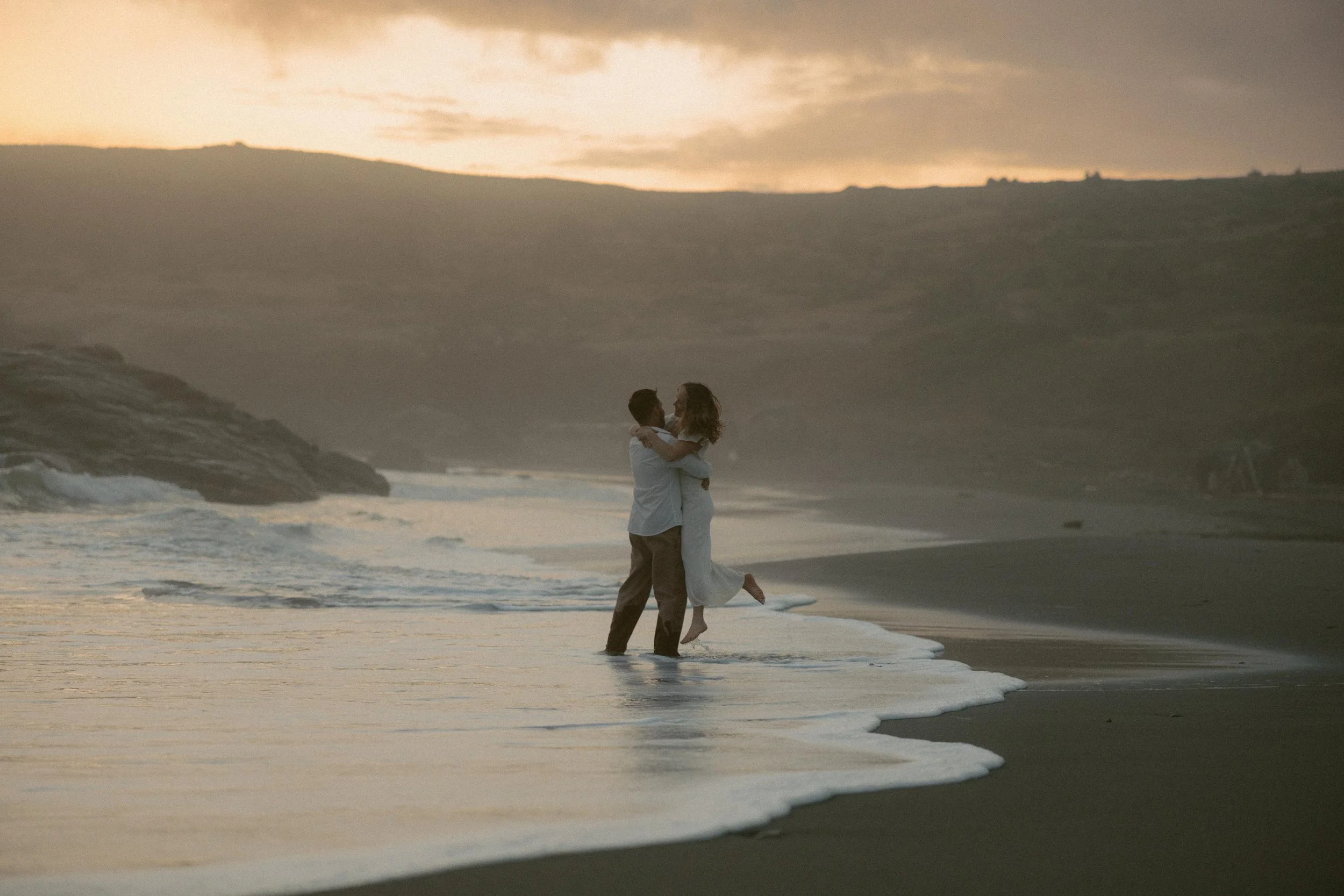 A couple is embracing and holding each other on the beach near the ocean waves during sunset or sunrise.