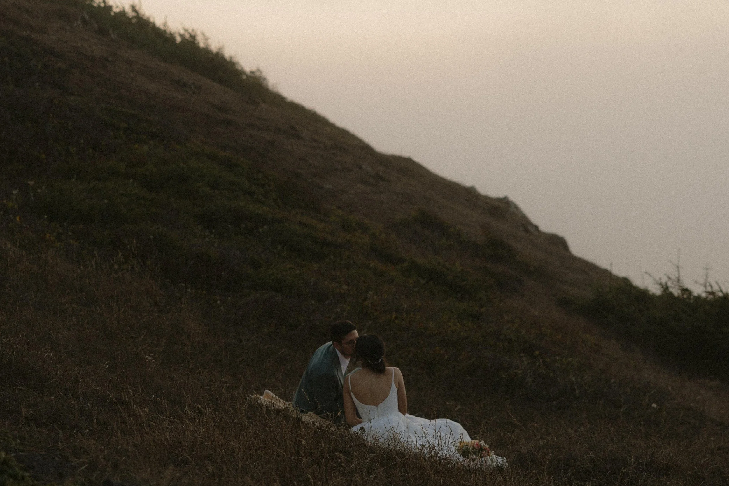 A couple sitting on the grass at the base of a hillside, embracing, with the sky overcast in the background.