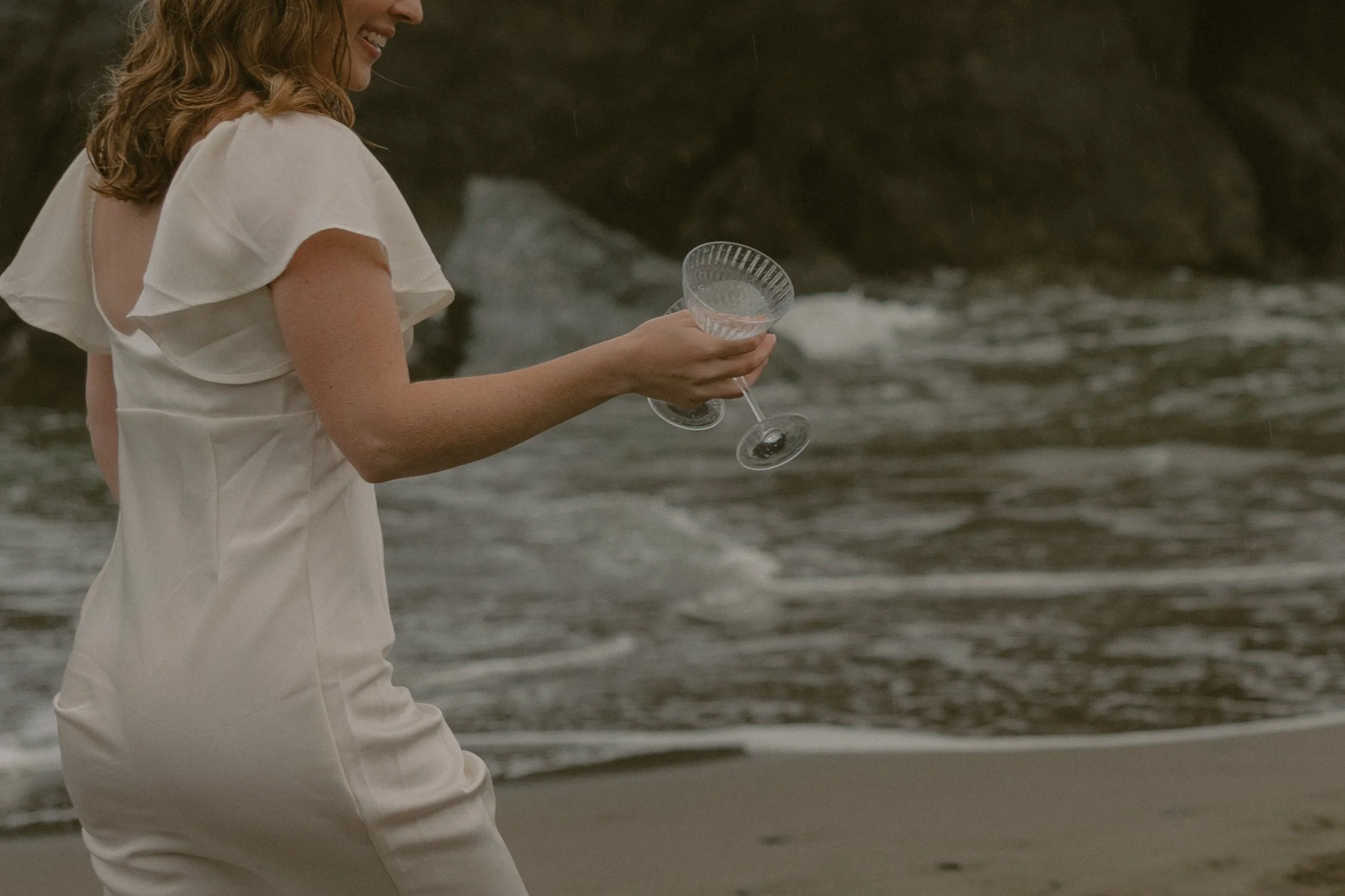 A woman in a white dress is holding an empty glass goblet, standing near the shoreline with waves in the background.