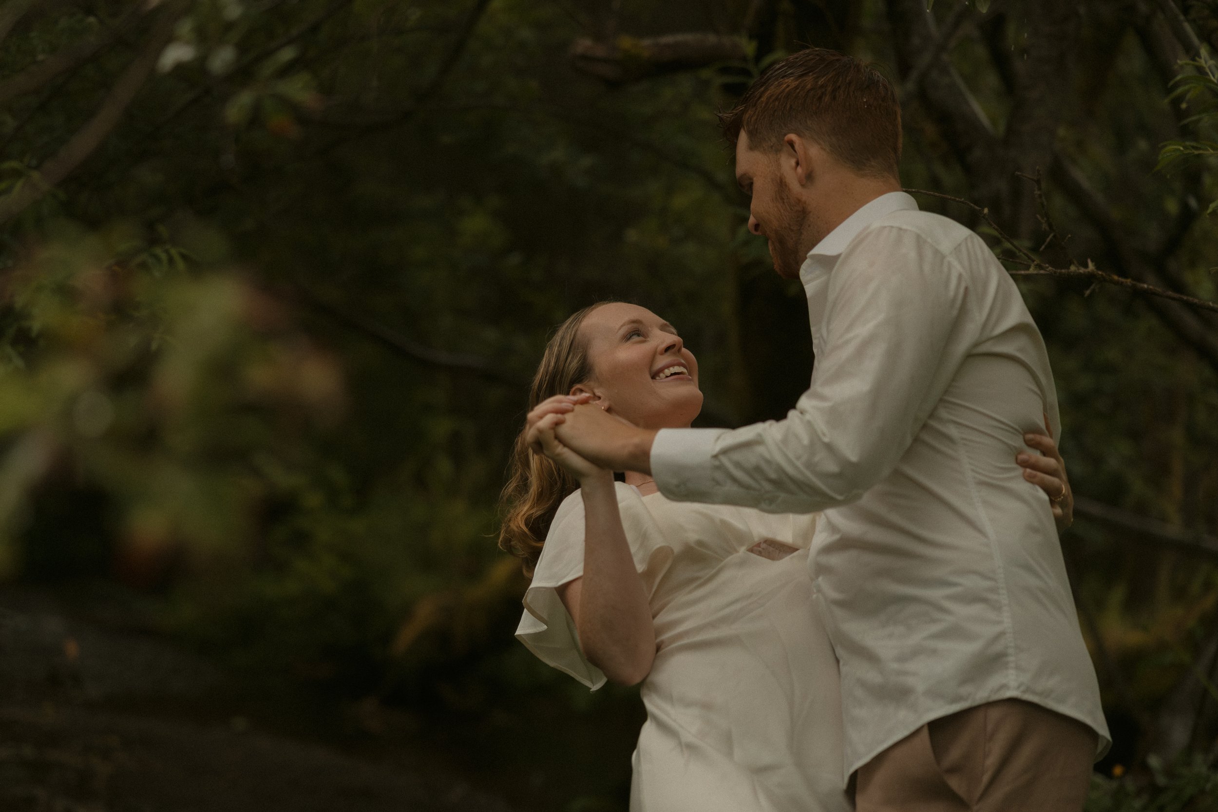 A couple dancing outdoors in a wooded area, smiling at each other.