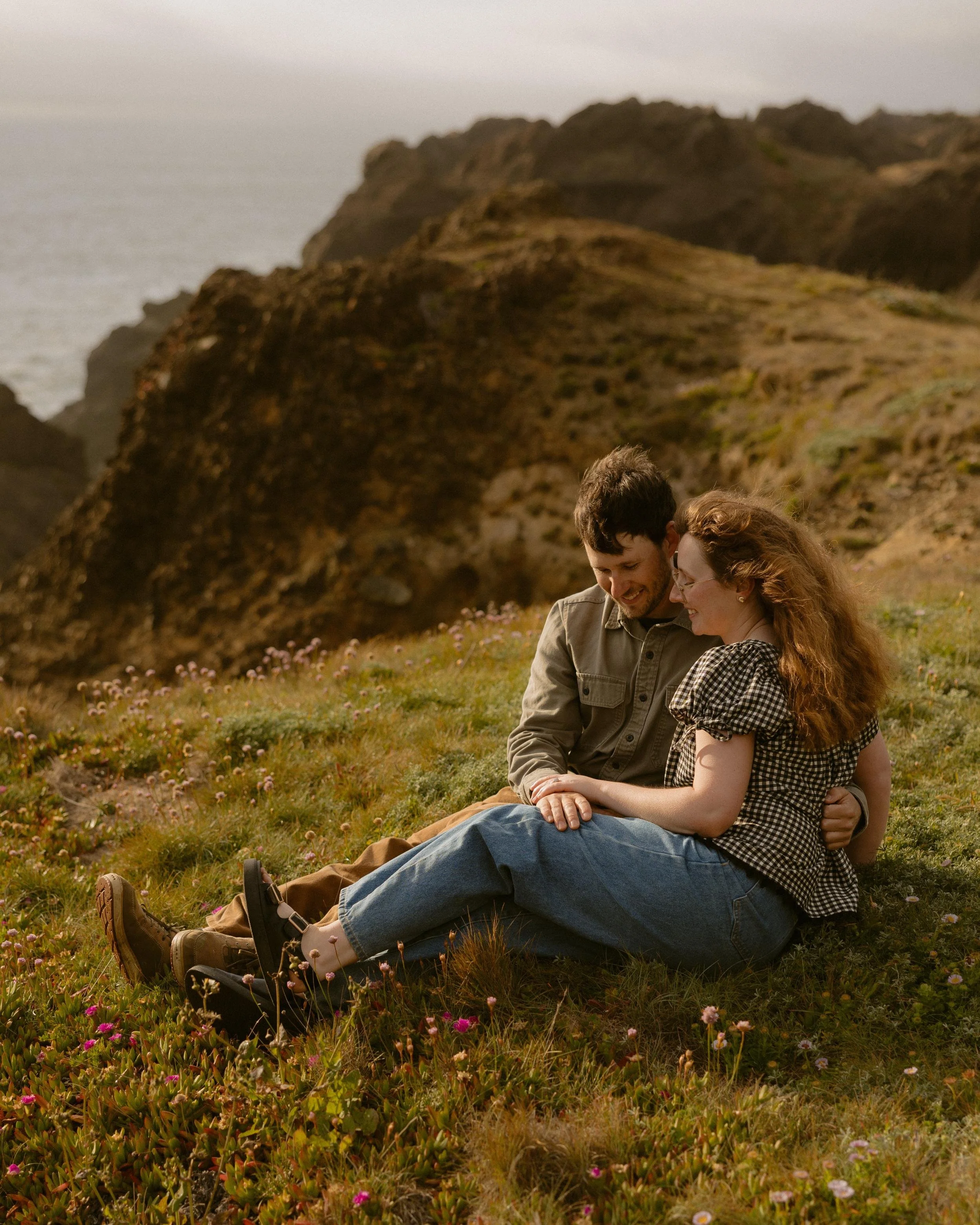 A couple sitting on grassy ground with pink and white flowers, outdoor landscape of hillside with large rocks, and cloudy sky in the background.