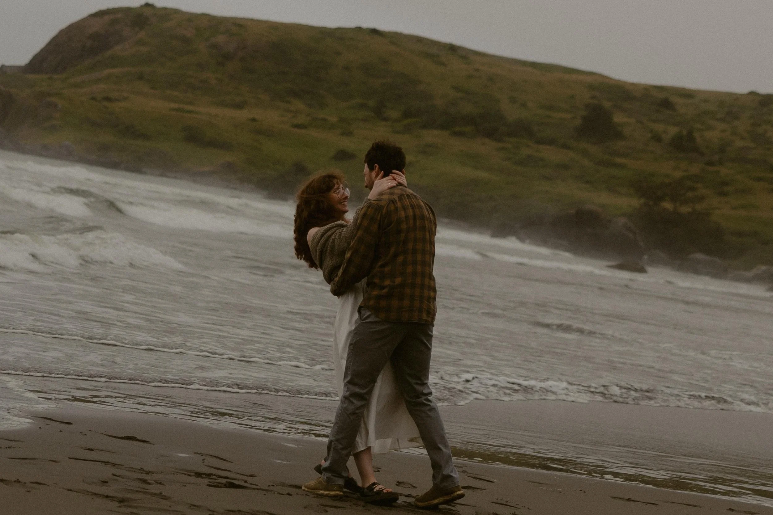 A couple standing on the beach near the ocean, embracing and looking at each other, with waves crashing nearby and a rocky hill in the background.