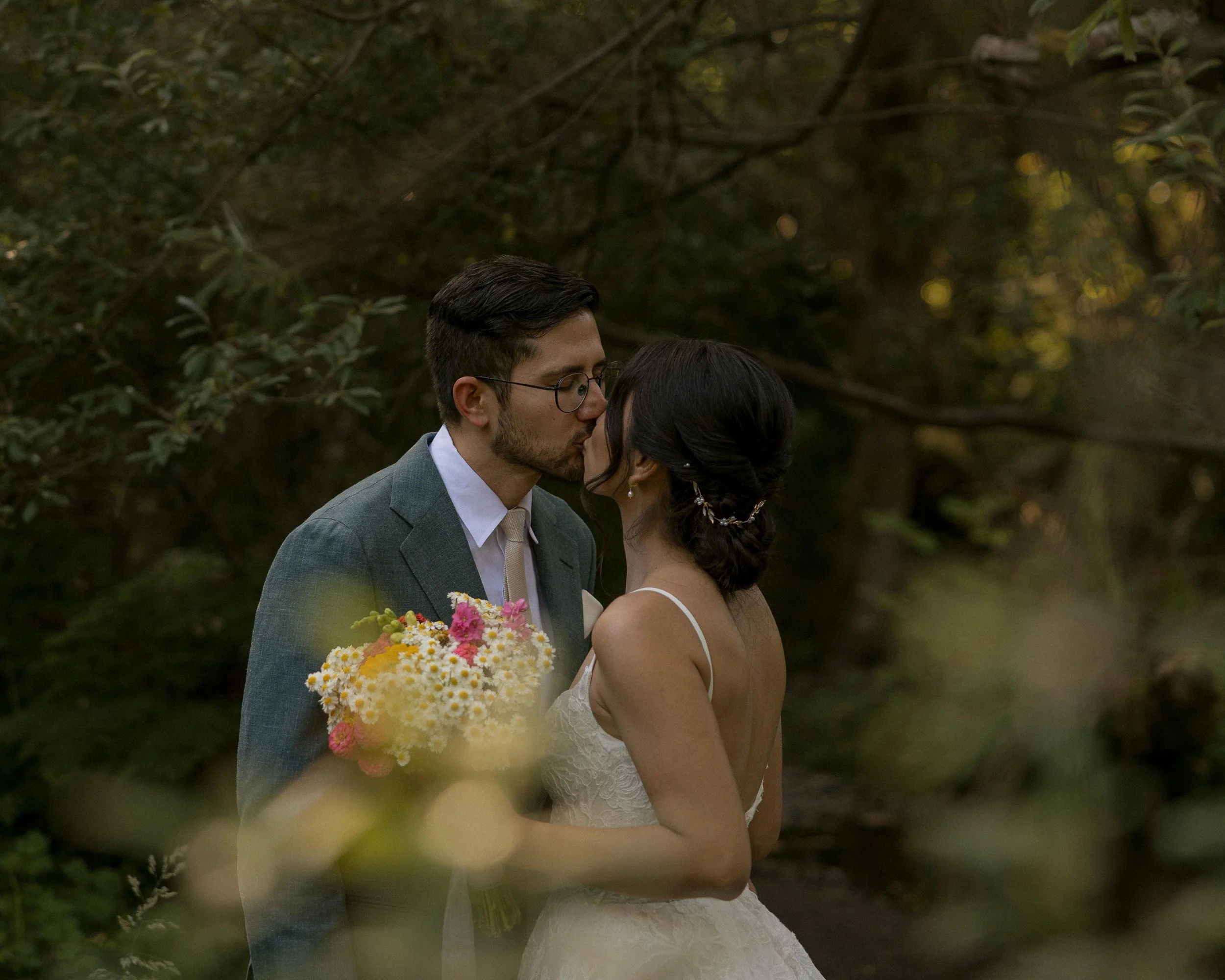 A couple dressed in wedding attire sharing a kiss outdoors during daylight, with the woman holding a bouquet of yellow and pink flowers, surrounded by greenery.