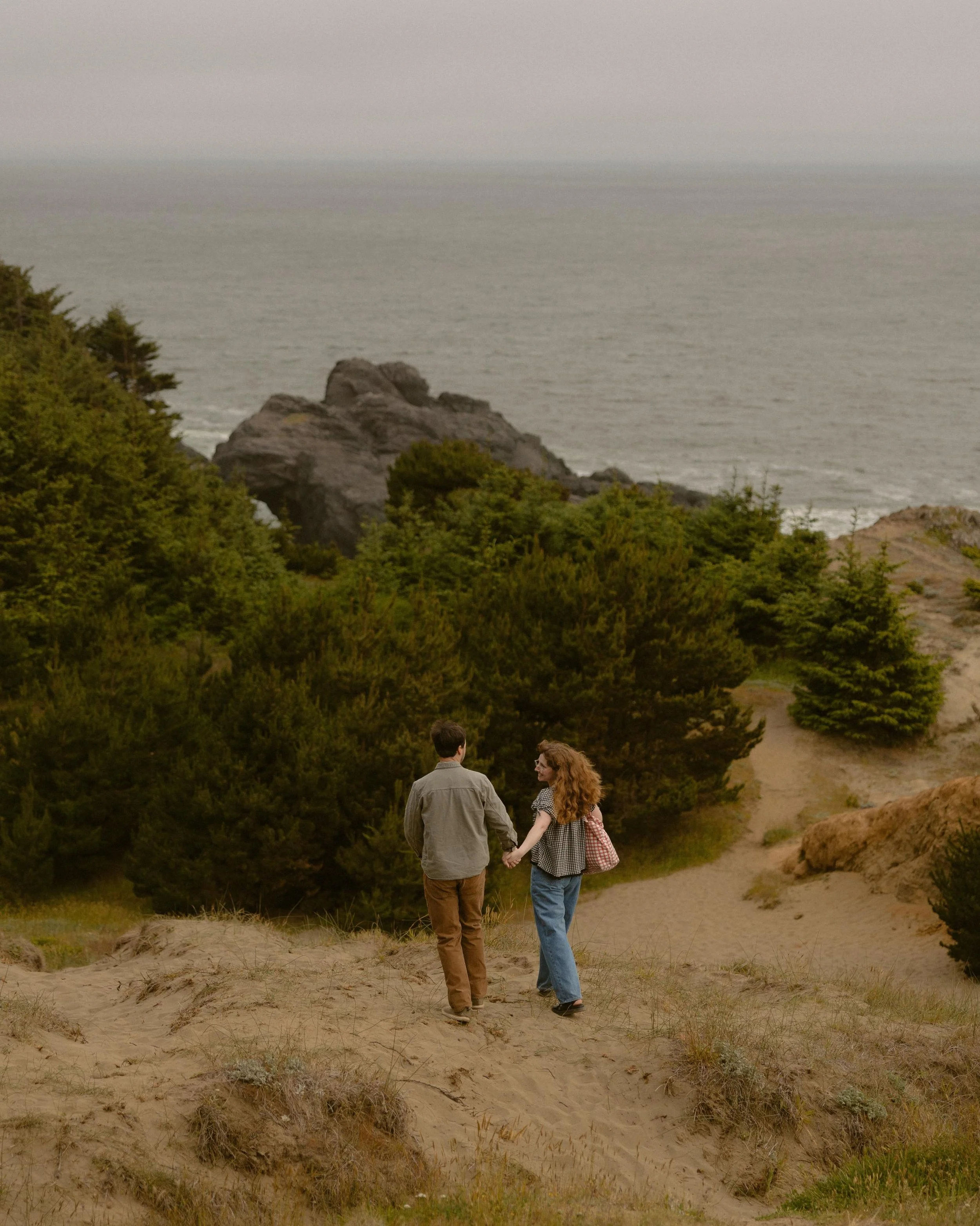 A couple holding hands and walking on a sandy beach trail with green bushes and trees, overlooking the ocean, during sunset or sunrise.