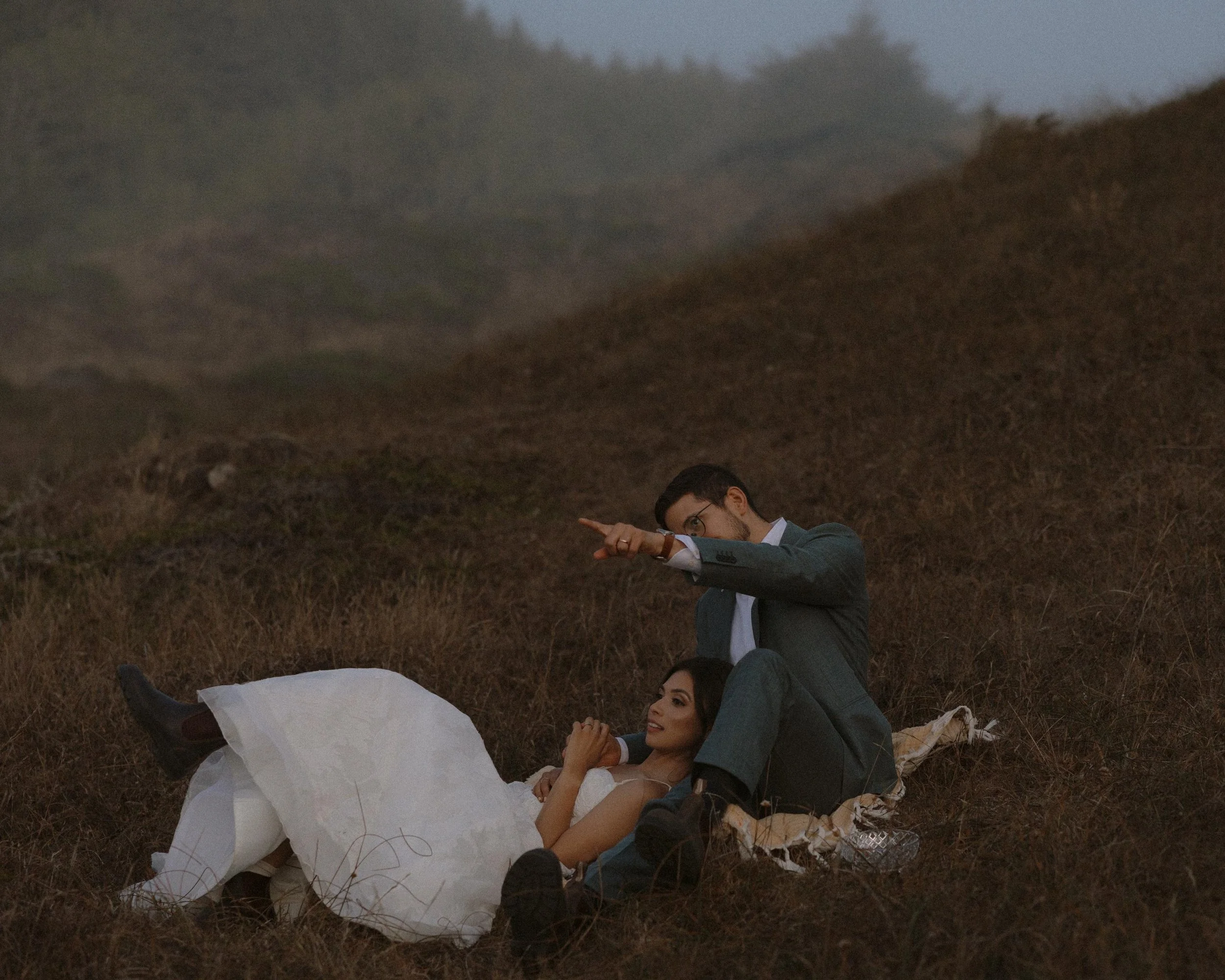 A man and woman lying on the ground outdoors at dusk, with the man pointing at something in the distance and the woman smiling.