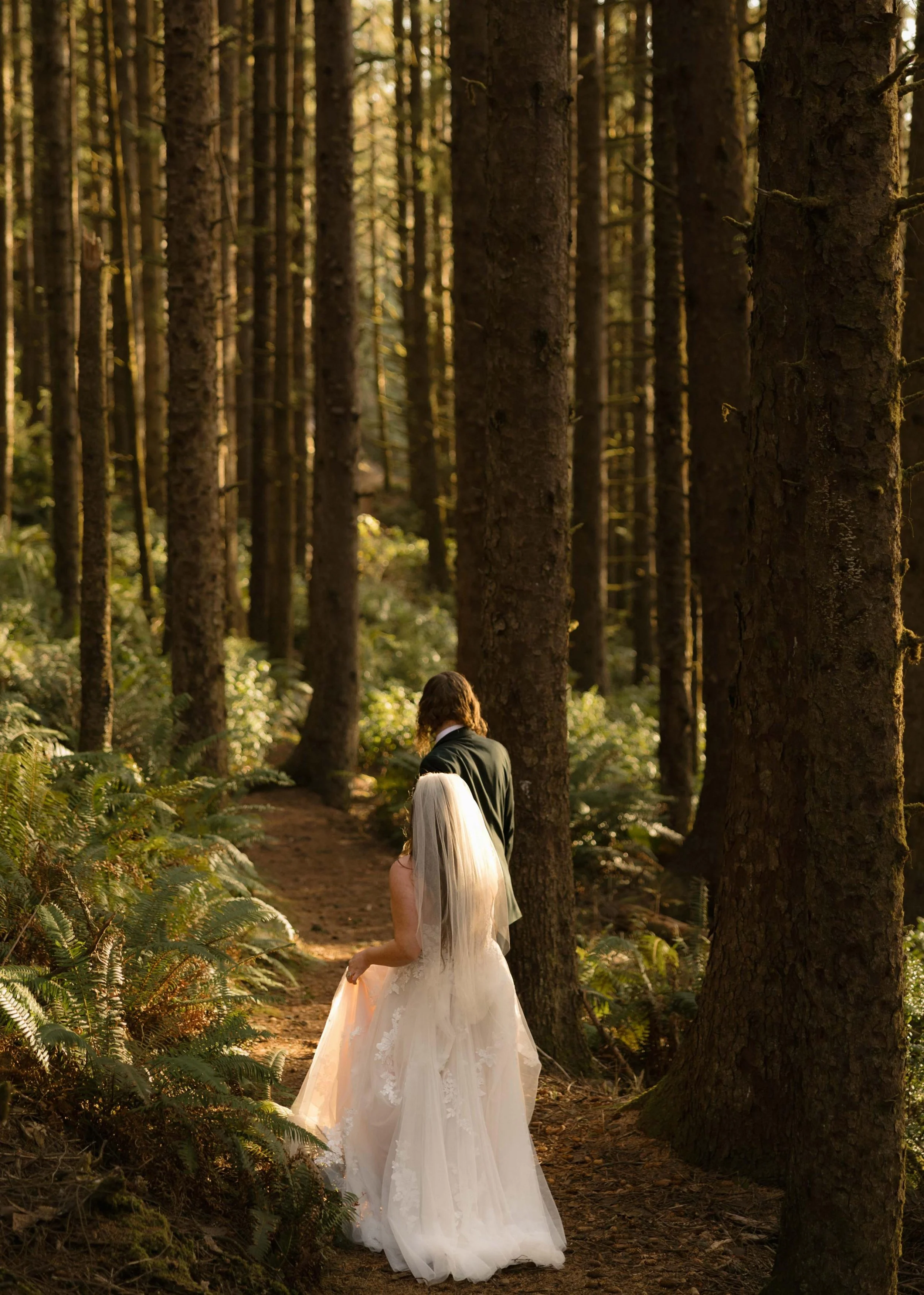 A bride in a white wedding dress and veil walking on a forest trail, with a man in a dark suit walking ahead of her.