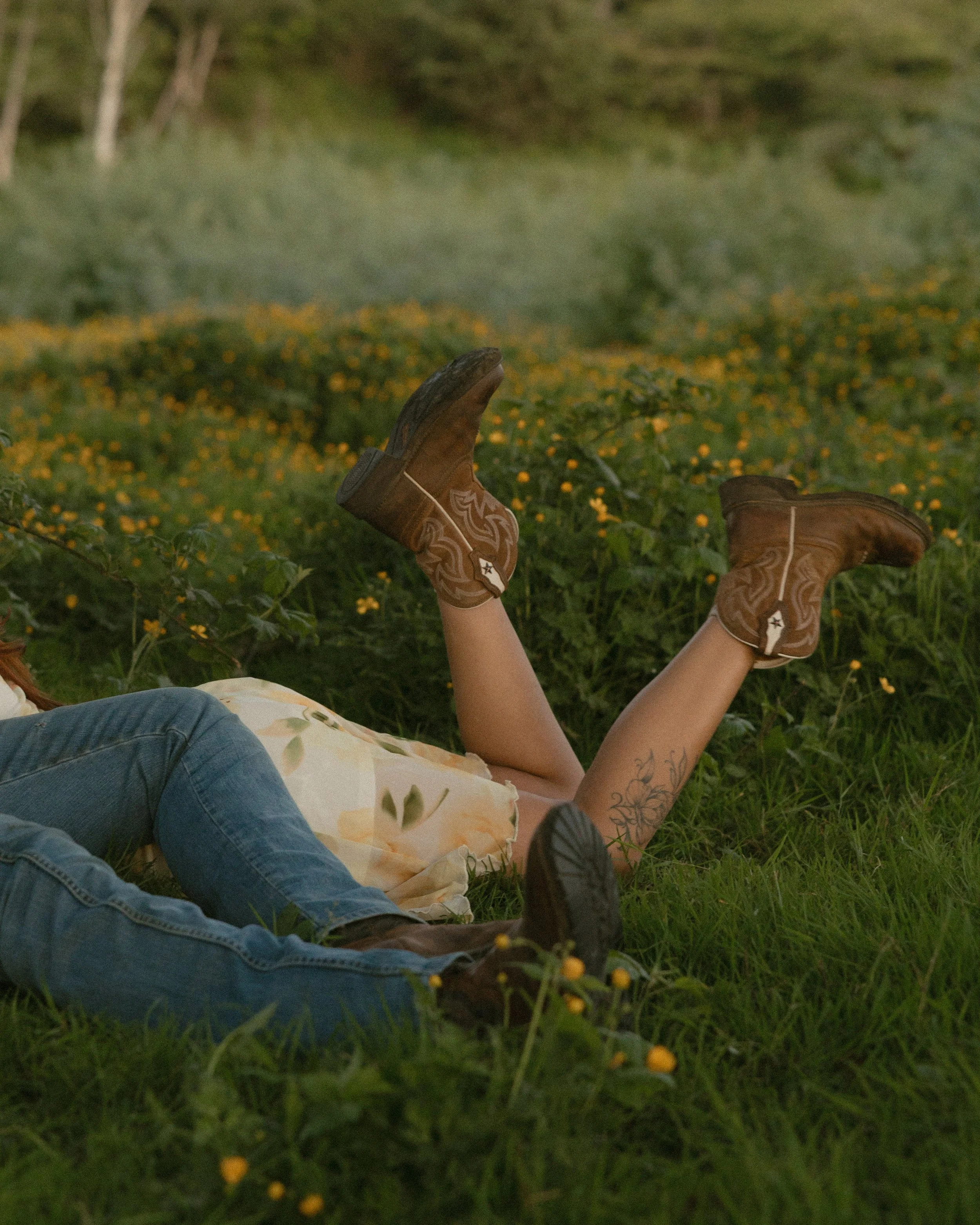 Person lying on grass with cowboy boots raised in the air, wearing jeans and a floral dress, surrounded by green foliage and small yellow flowers.