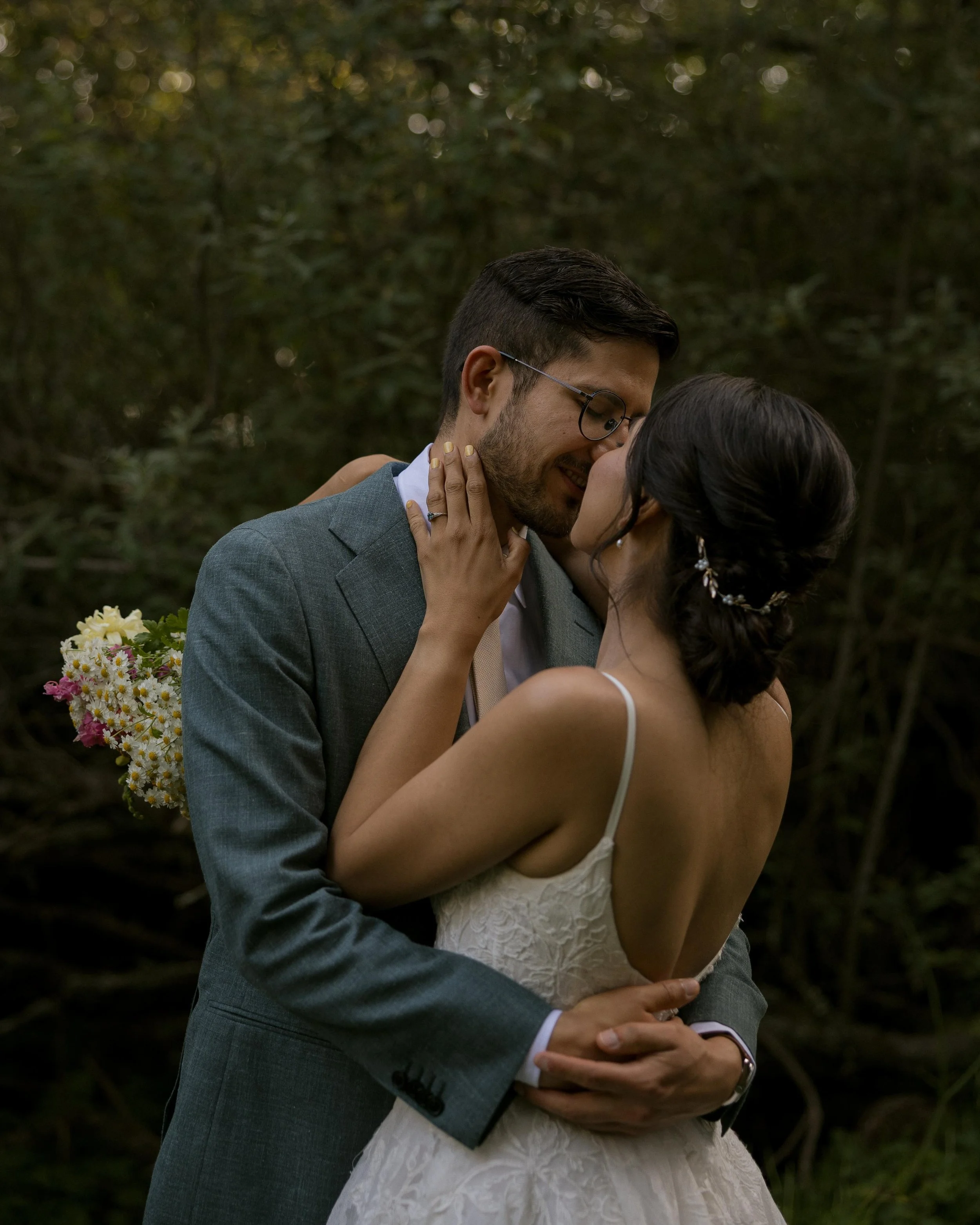A couple in wedding attire embracing outdoors, with the groom wearing a gray suit and glasses, and the bride in a white dress with lace details. They are close, with their faces touching, against a backdrop of greenery and soft lighting.