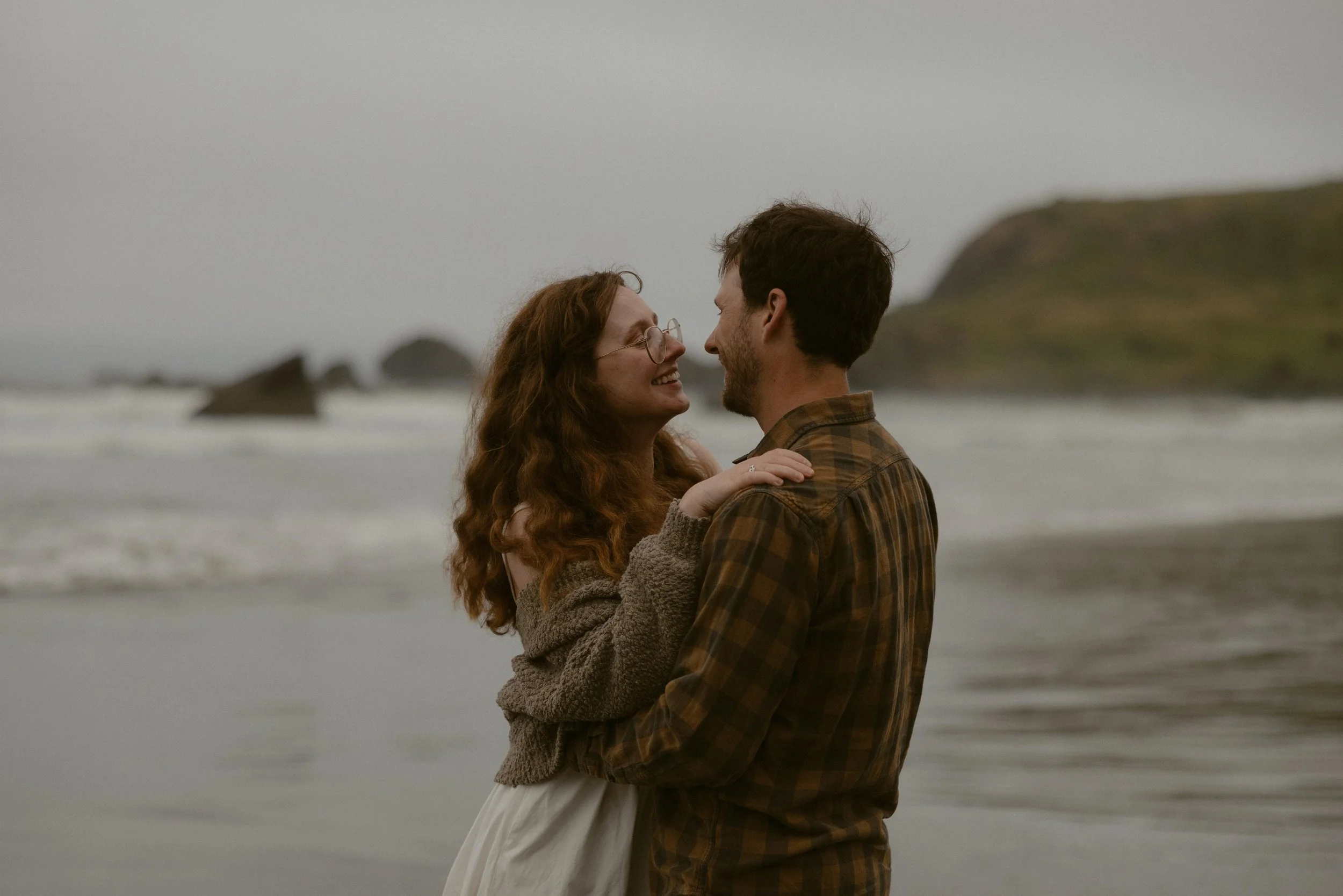 A couple embracing and smiling at each other on a beach with rocks and hills in the background.
