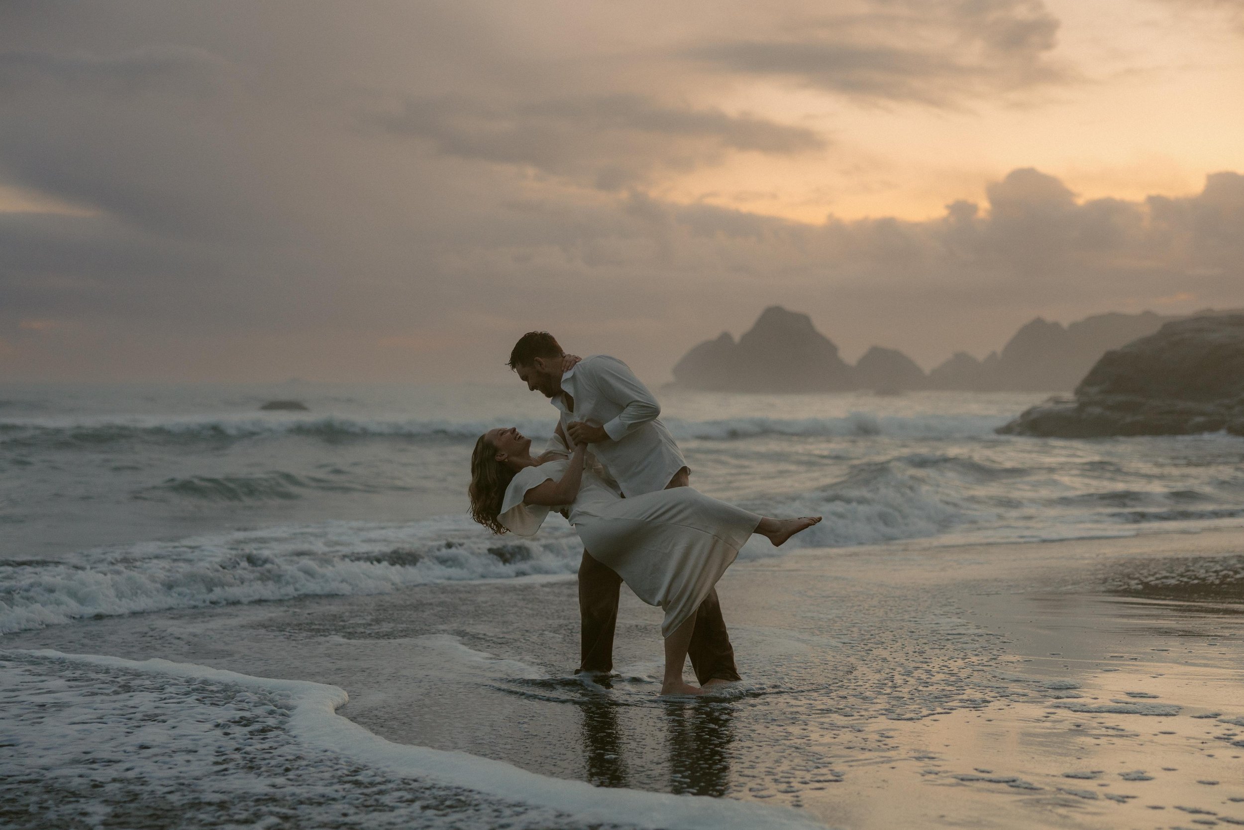 A couple in wedding attire dancing in the ocean at sunset, with rocky islands in the background.