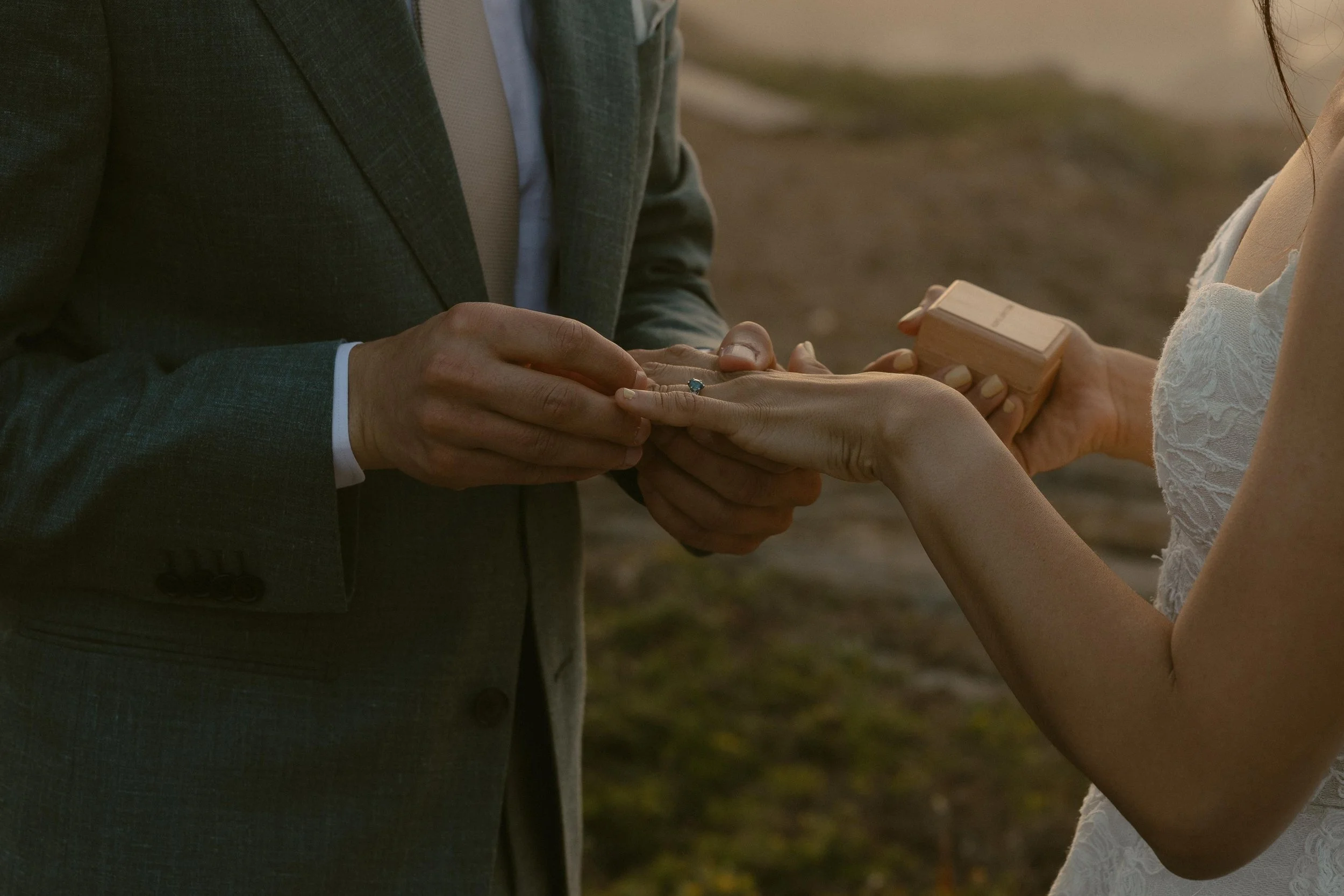 A close-up of a marriage proposal where a man in a gray suit is holding a woman's hand, revealing a ring with a large blue gemstone. The woman, dressed in white lace, is receiving the ring with a small wooden box in her other hand. The scene is outdo