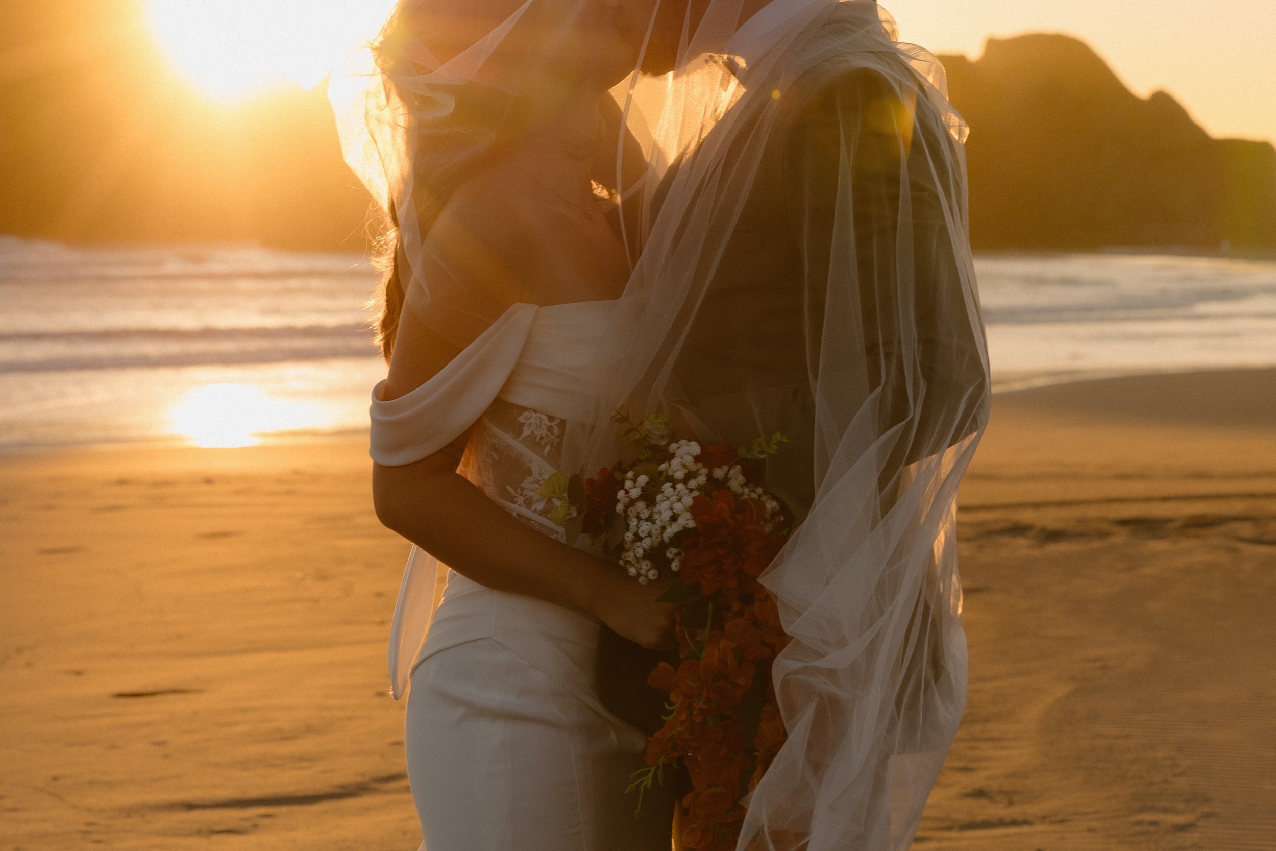 A couple dressed in wedding attire embracing on the beach at sunset, holding a bouquet of flowers.