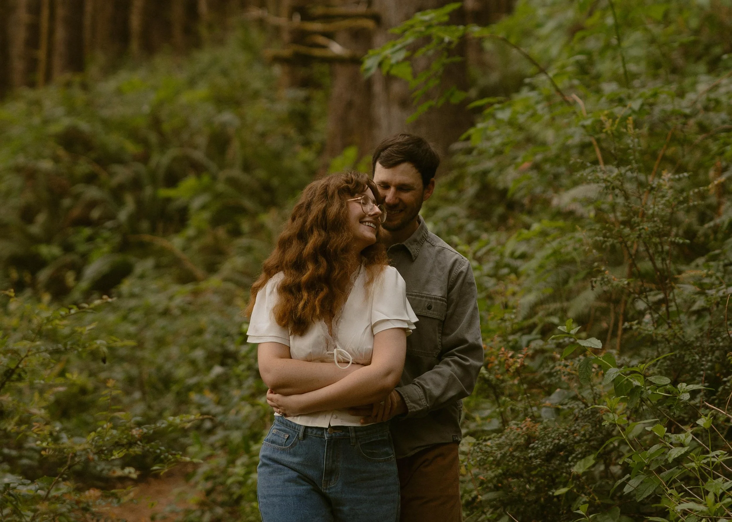 A couple smiling and embracing each other in a forest with green foliage.
