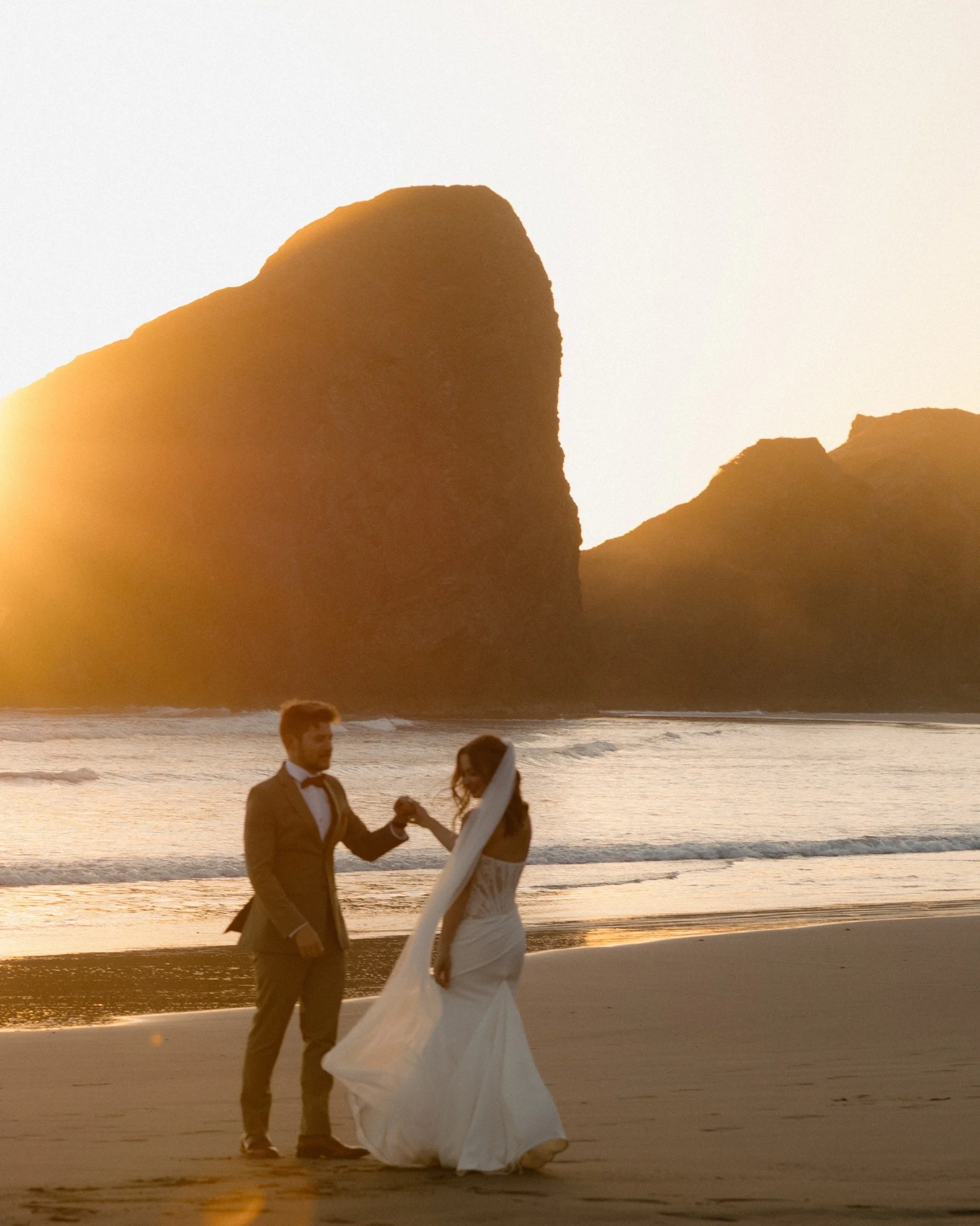A man and woman in wedding attire holding hands on a beach at sunset, with large rock formations and a surfboard between them.
