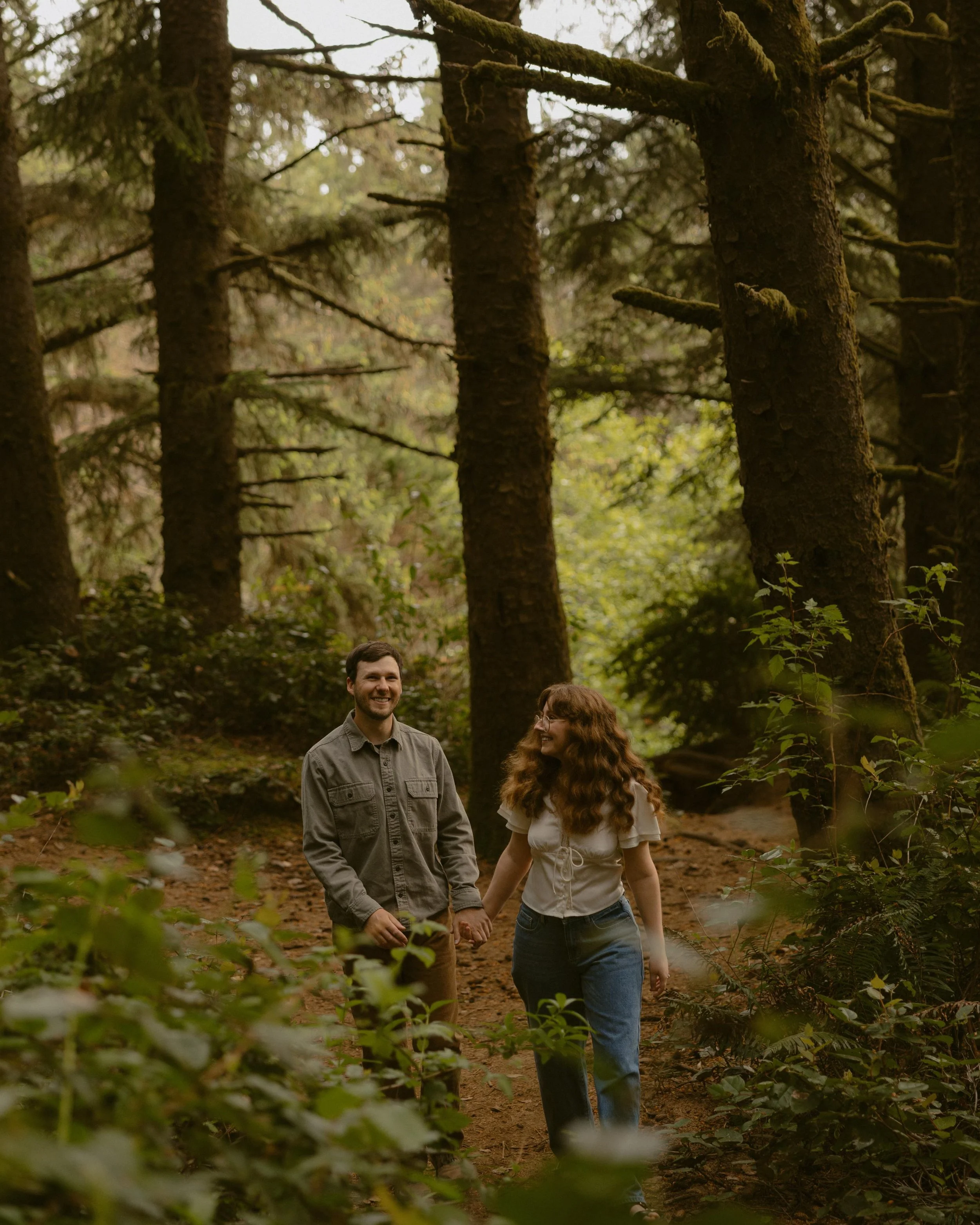 A young man and woman walking hand in hand through a forest, smiling and enjoying nature on a cloudy day.