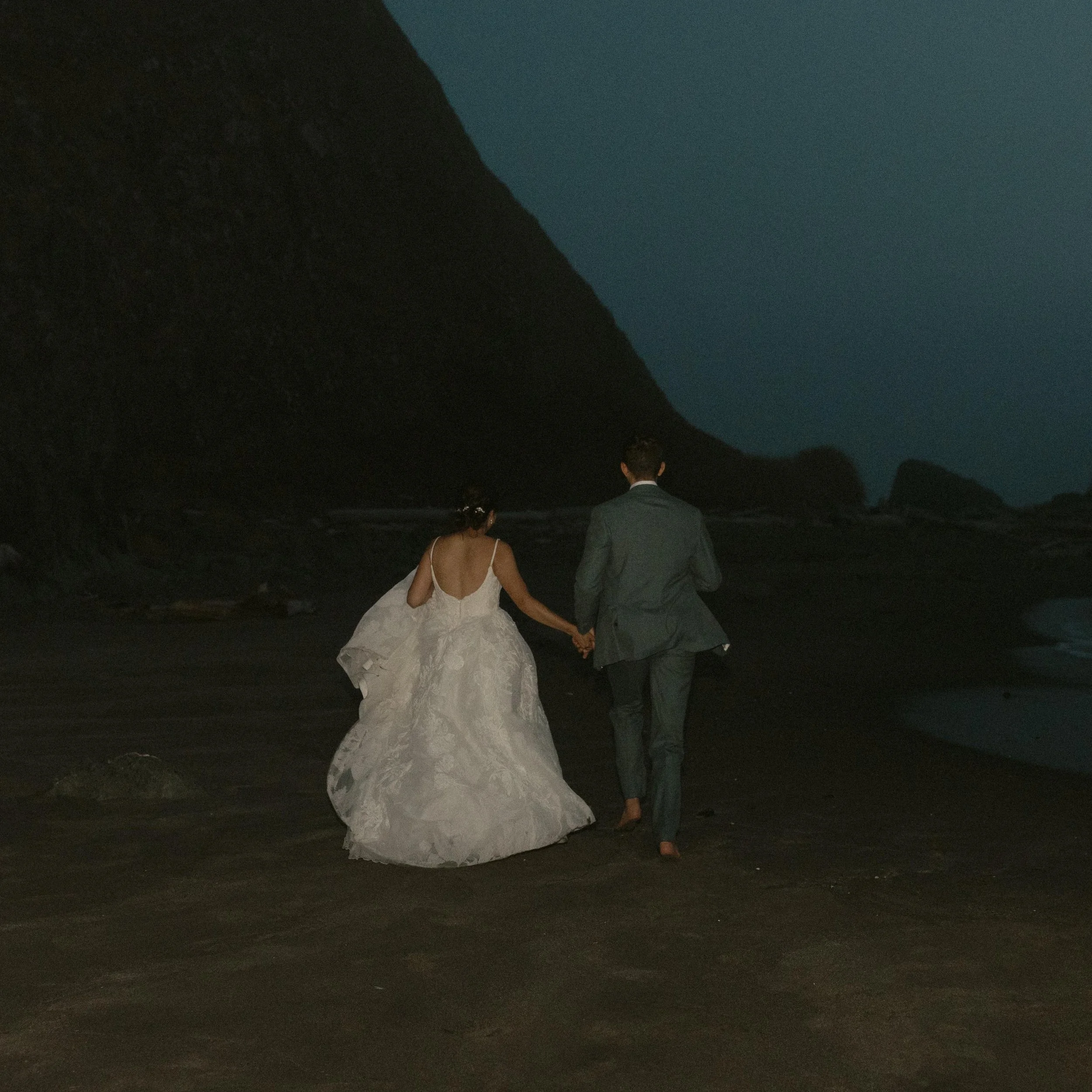A bride and groom walking hand in hand on a beach at night, with rocky cliffs in the background and the ocean ahead.