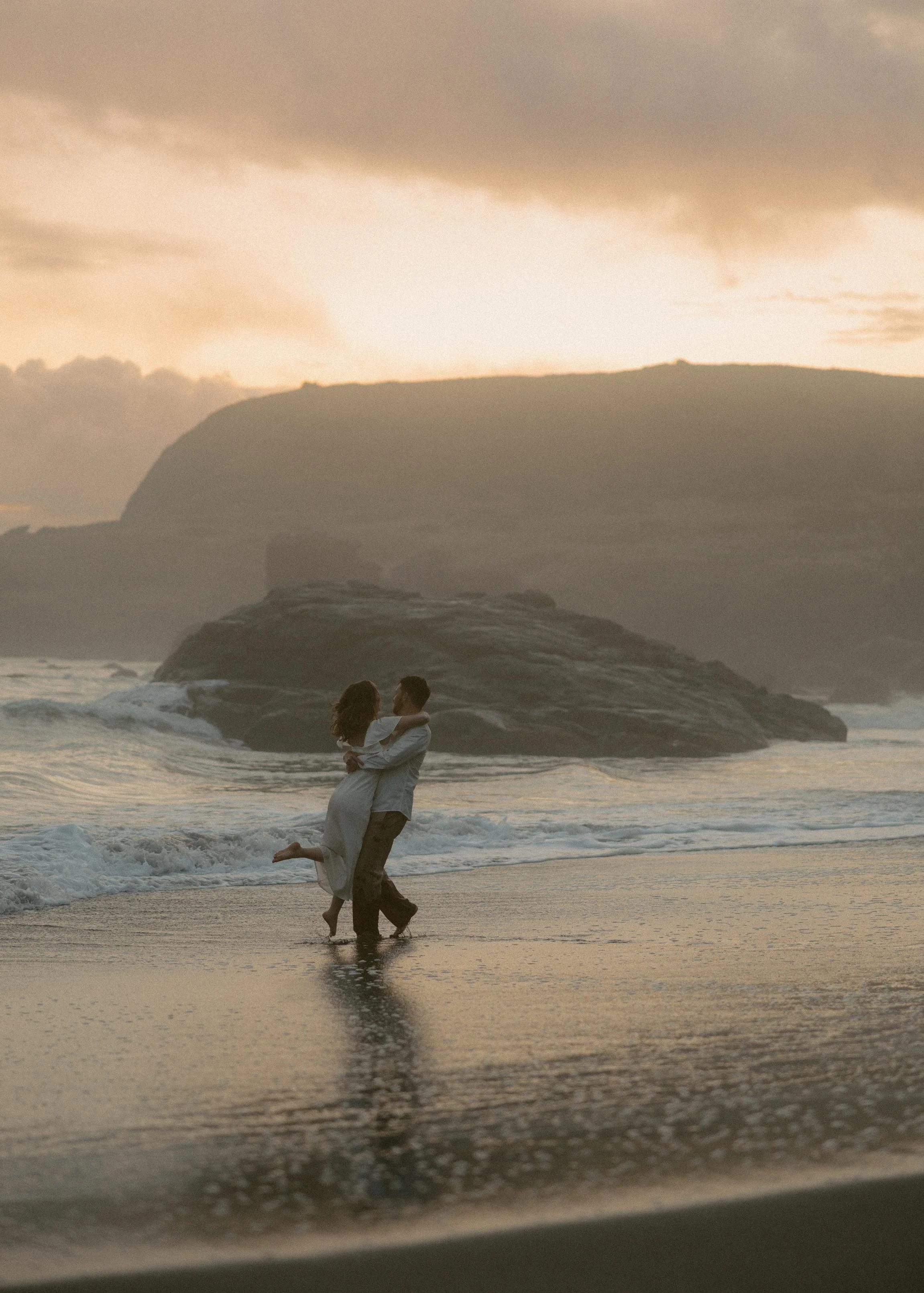 A couple is at the beach, with one person carrying the other in their arms. The scene is at sunset with the sky partly cloudy and large rocks in the background, and the ocean waves gently crashing onto the shore.