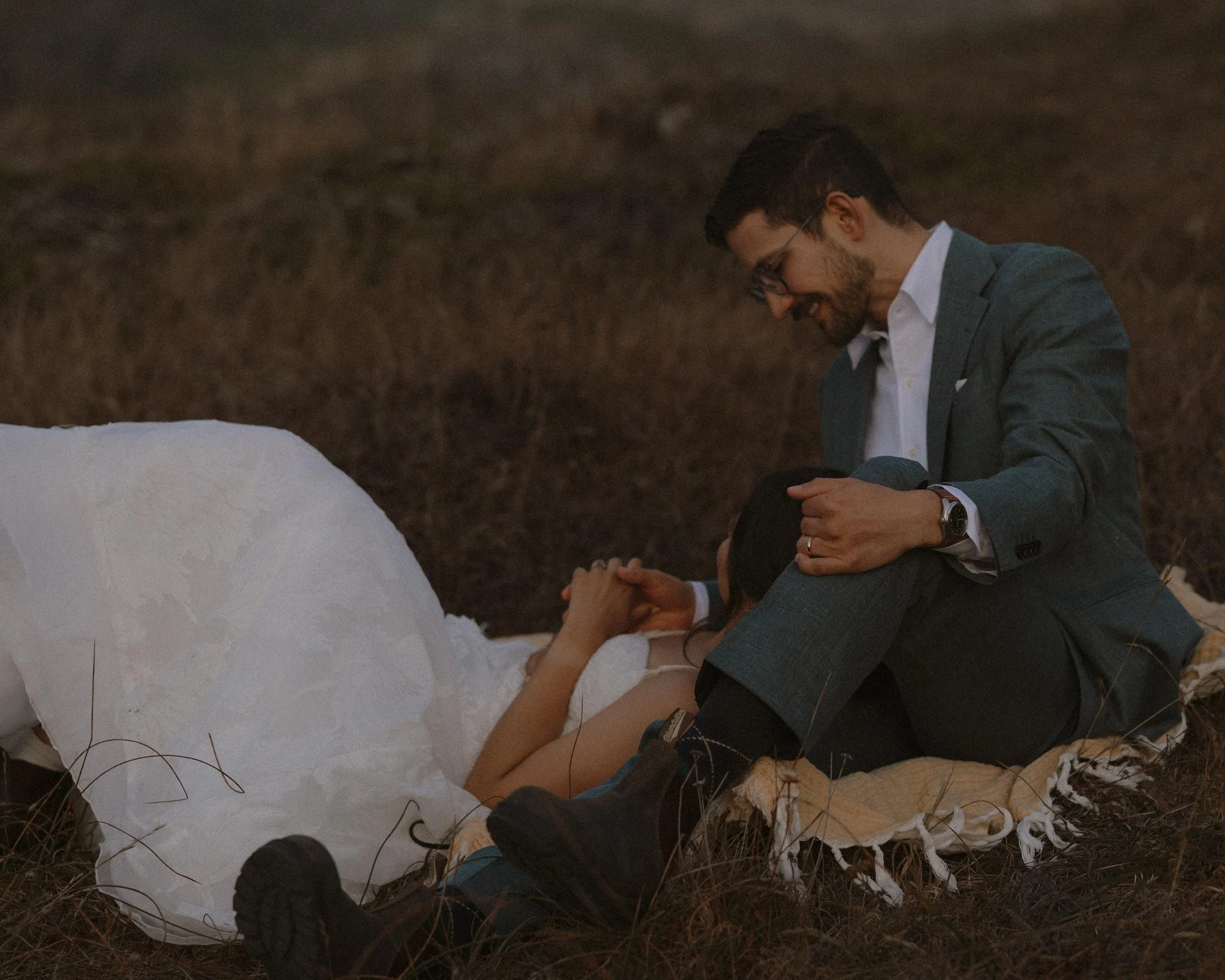 A man wearing glasses in a suit, smiling, and sitting on the ground next to a woman lying on her back. The woman is wearing a white dress, and her hands are clasped together. They are outdoors on a rocky and grassy terrain with some dry grass in the 