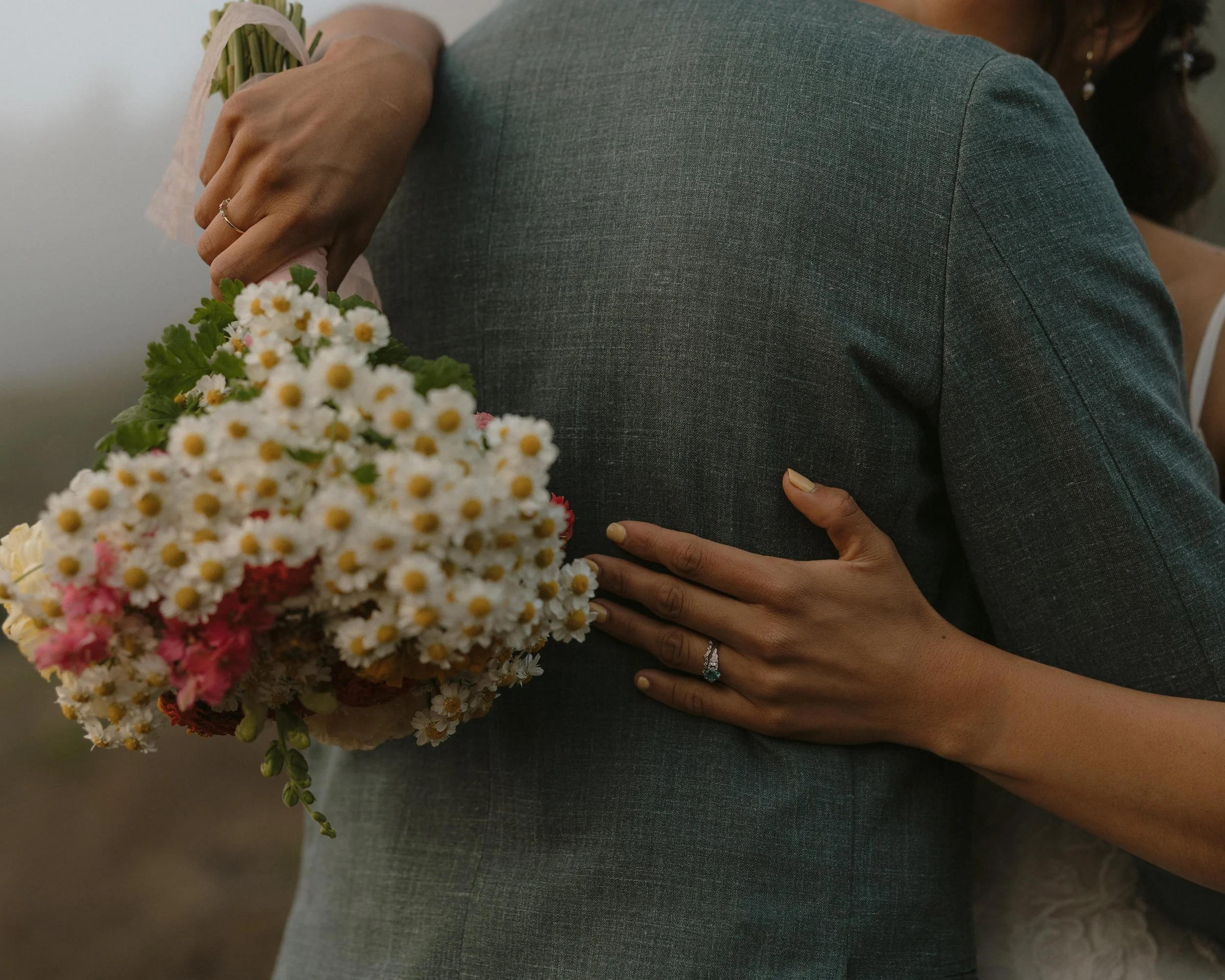A person holding a bouquet of white and pink flowers, wearing a gray blazer, with one hand resting on their back and the other holding the bouquet close to their chest.