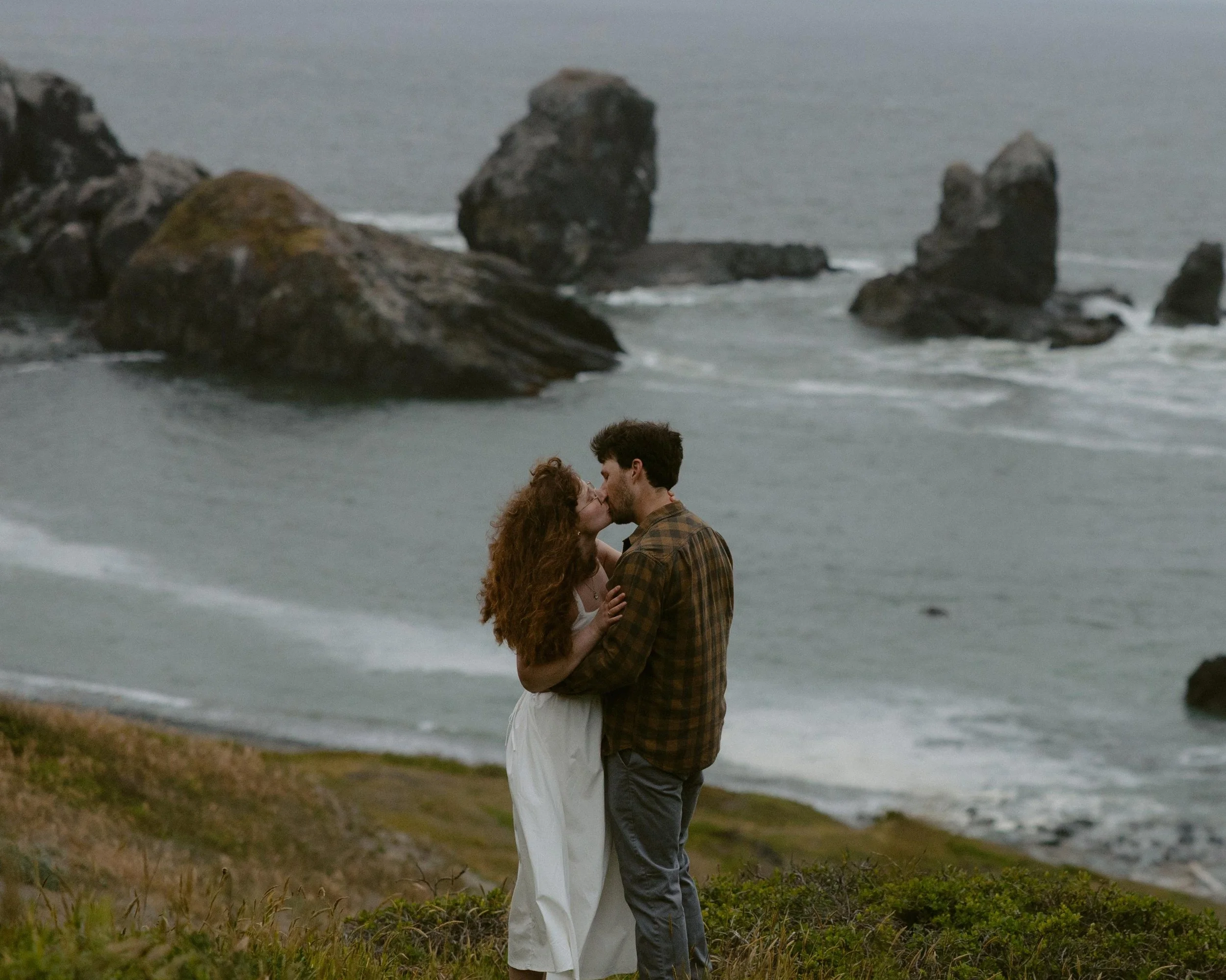 A couple stands close together on a grassy area near the beach, sharing a kiss as waves crash against rocks in the background.
