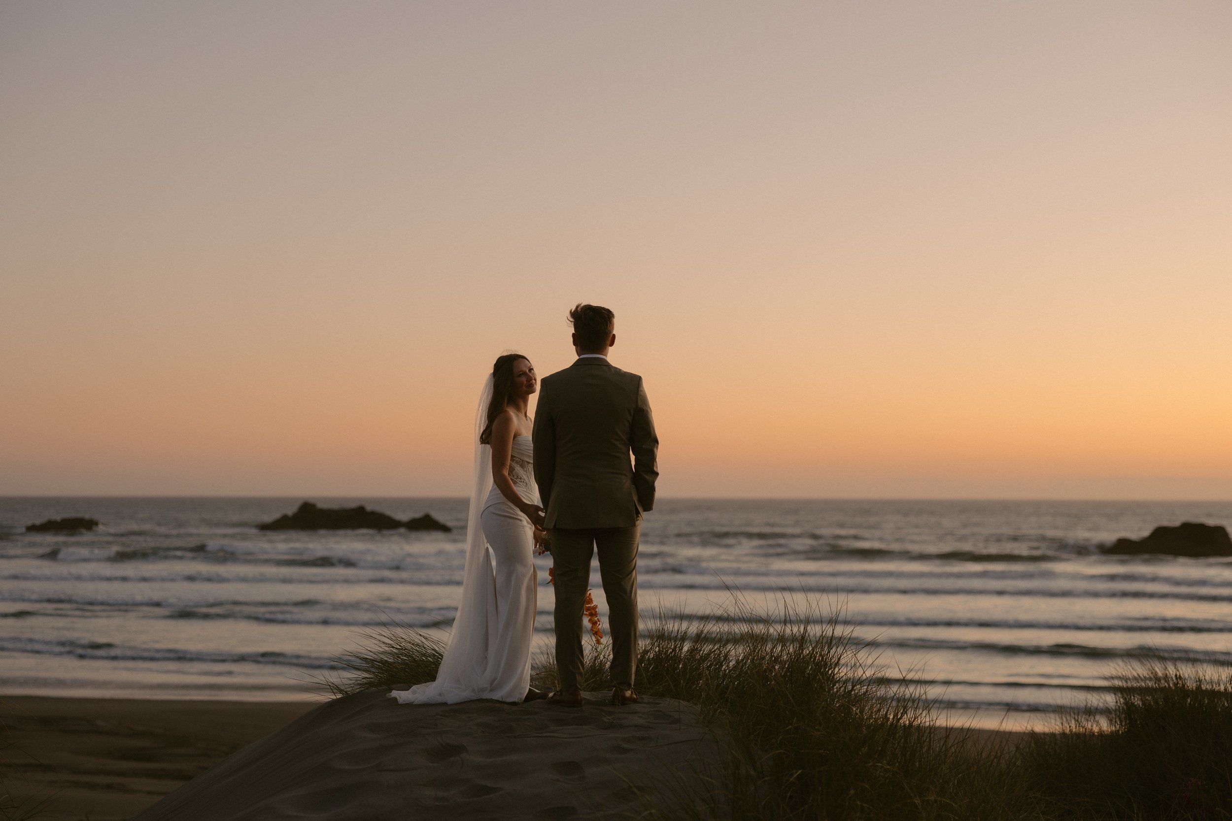 A bride and groom holding hands on the beach during sunset, facing the ocean, with rocks and grass in the foreground.