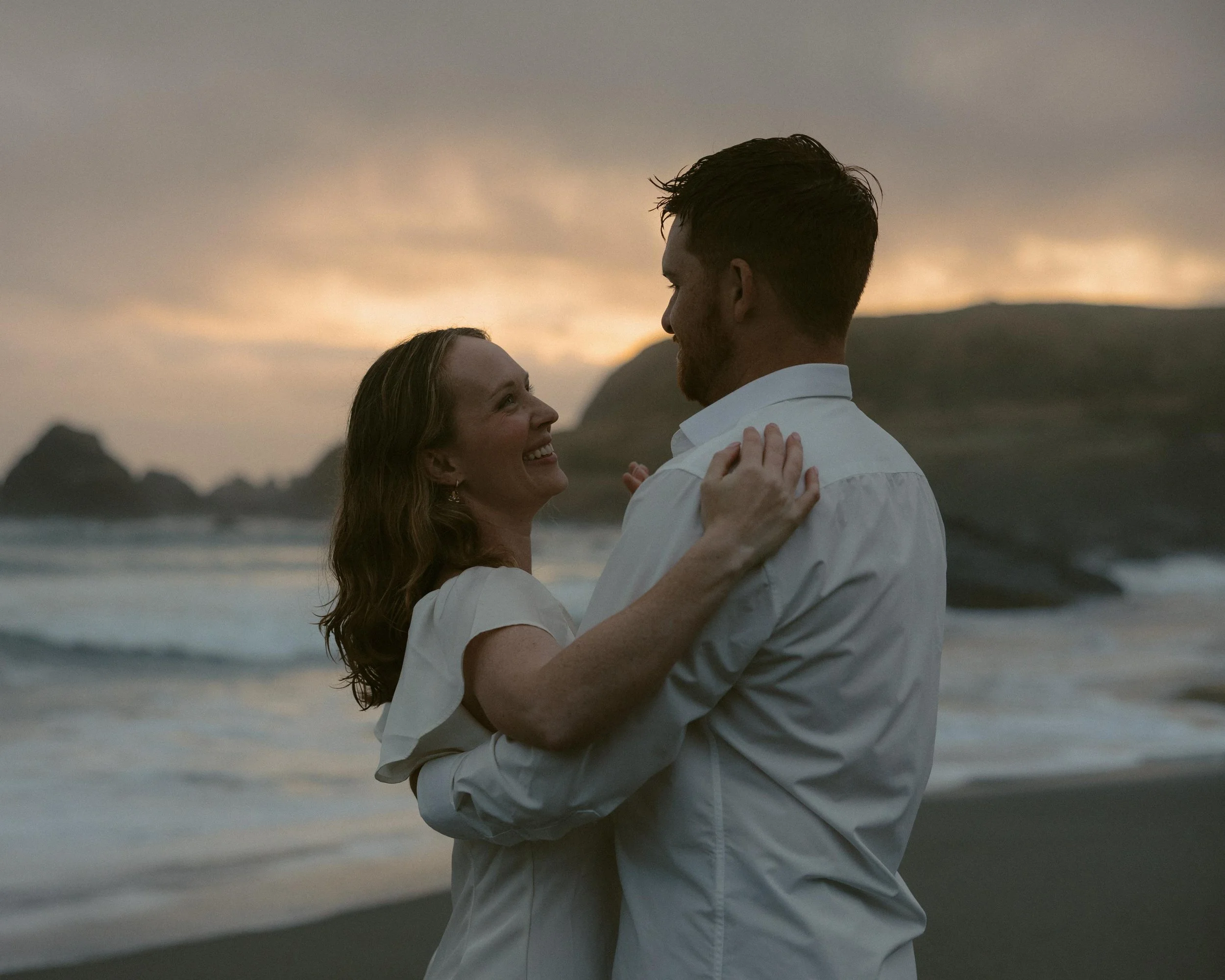 A couple dancing and smiling at the beach during sunset, with mountains in the background.