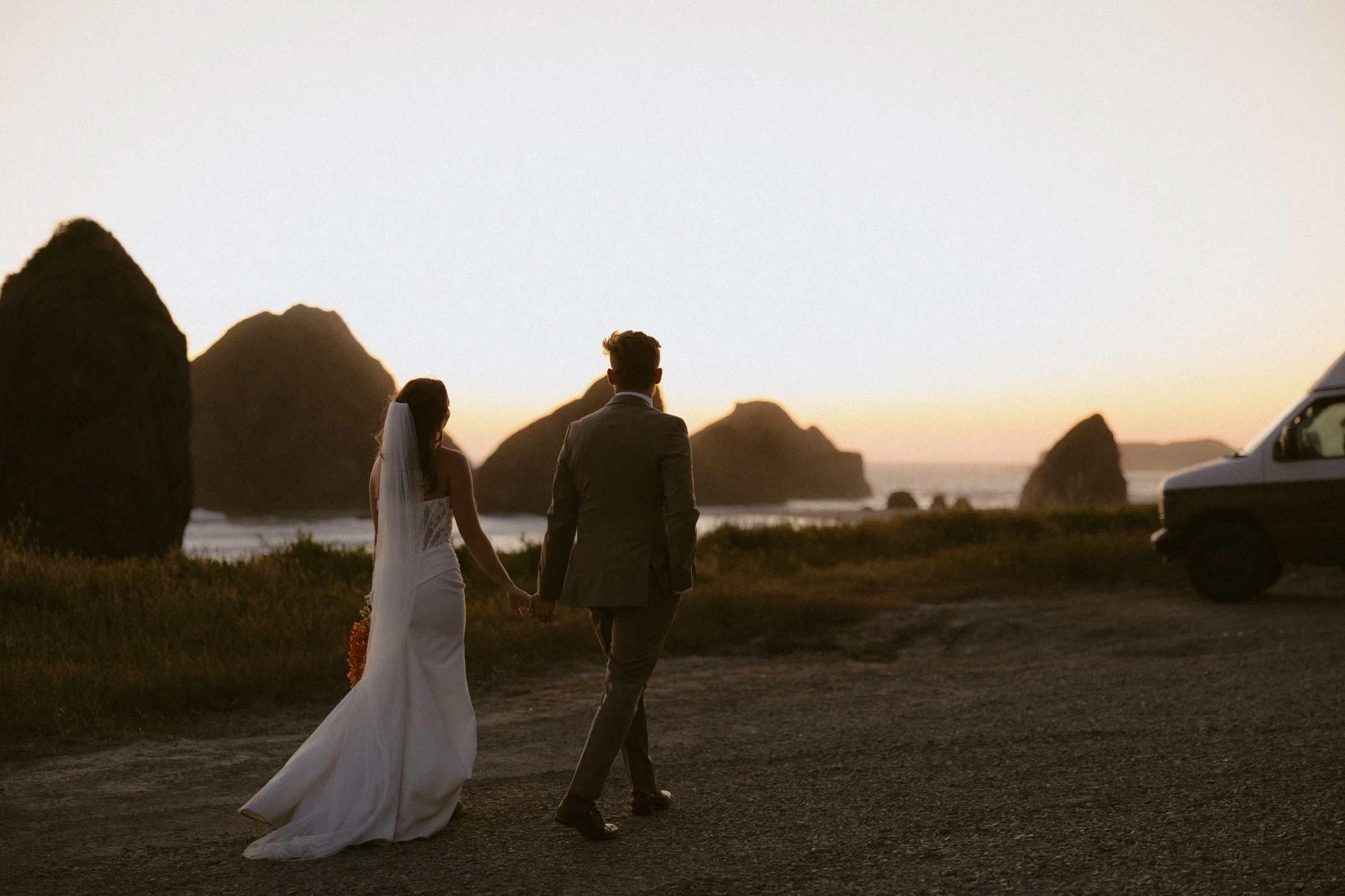 A couple in wedding attire walking hand in hand on a beach during sunset with large rocks in the background.