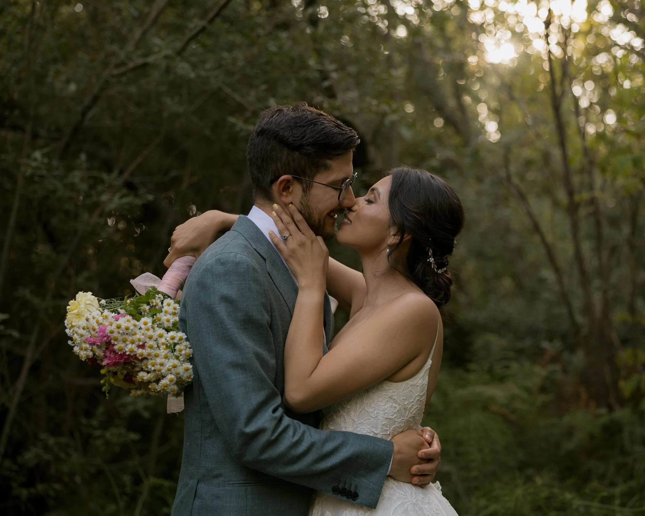 A couple in wedding attire sharing an intimate moment outdoors, with the man in a gray suit and glasses and the woman in a white lace wedding dress, holding a bouquet of colorful flowers, surrounded by trees and warm sunlight.