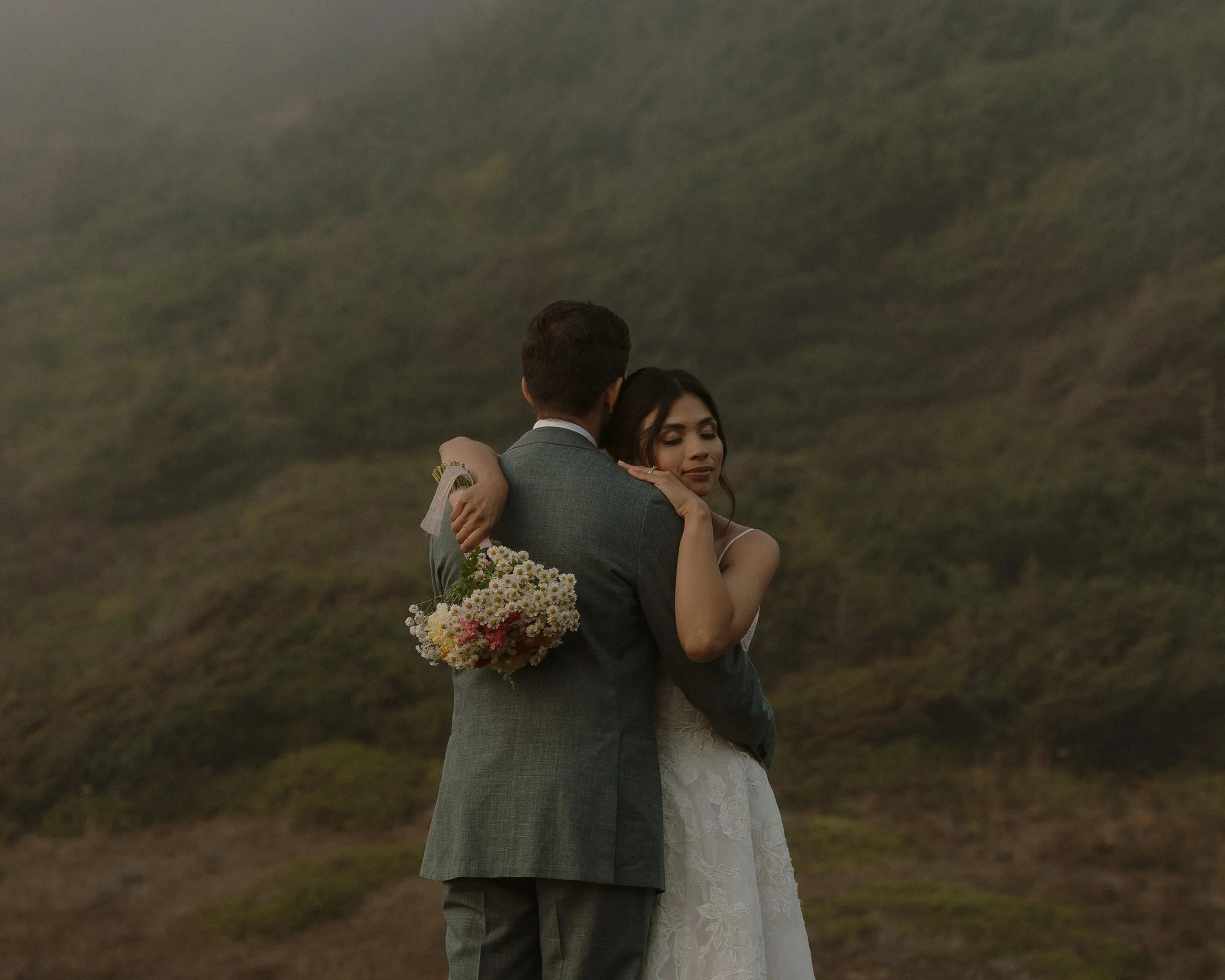 A couple embraces outdoors in wedding attire with a woman holding a bouquet of flowers, hills or mountains in the background.