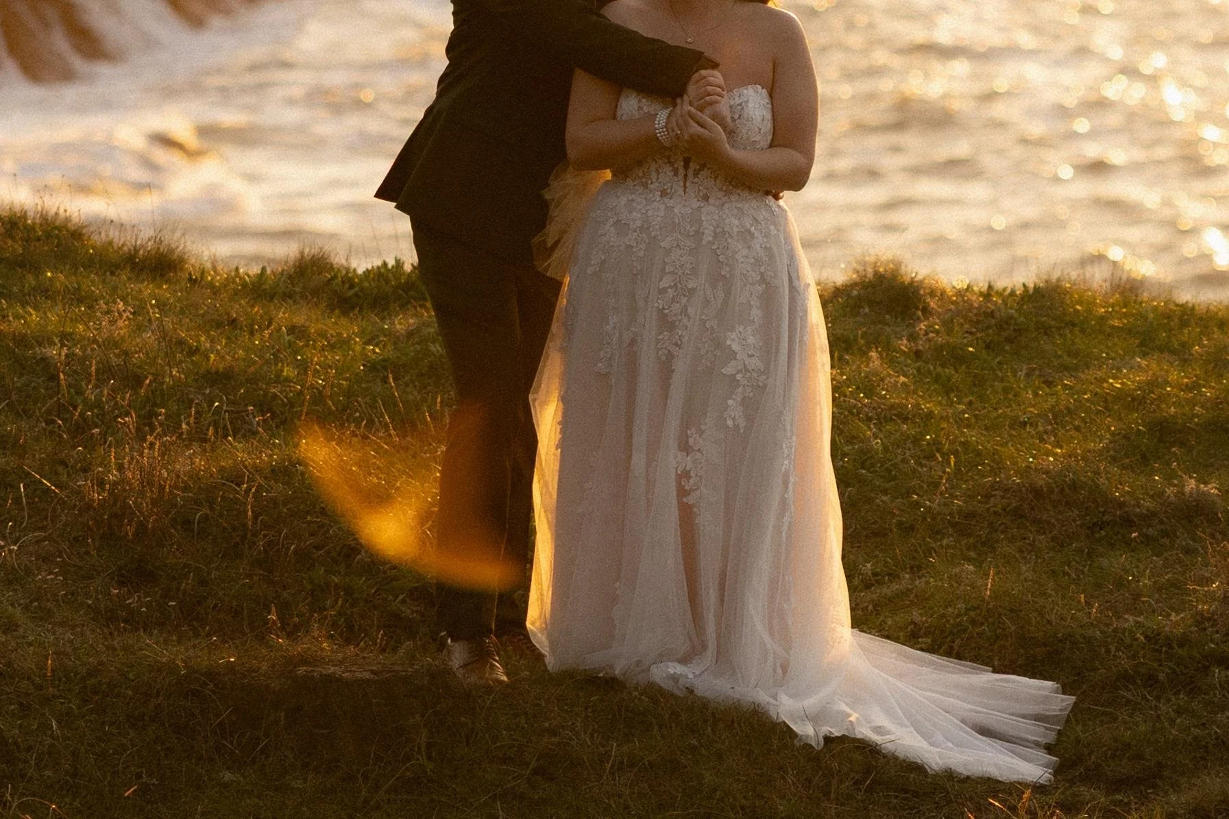 A bride in a lace wedding dress standing outdoors on grass near the water during sunset, with a man in a dark suit beside her, holding her hand.