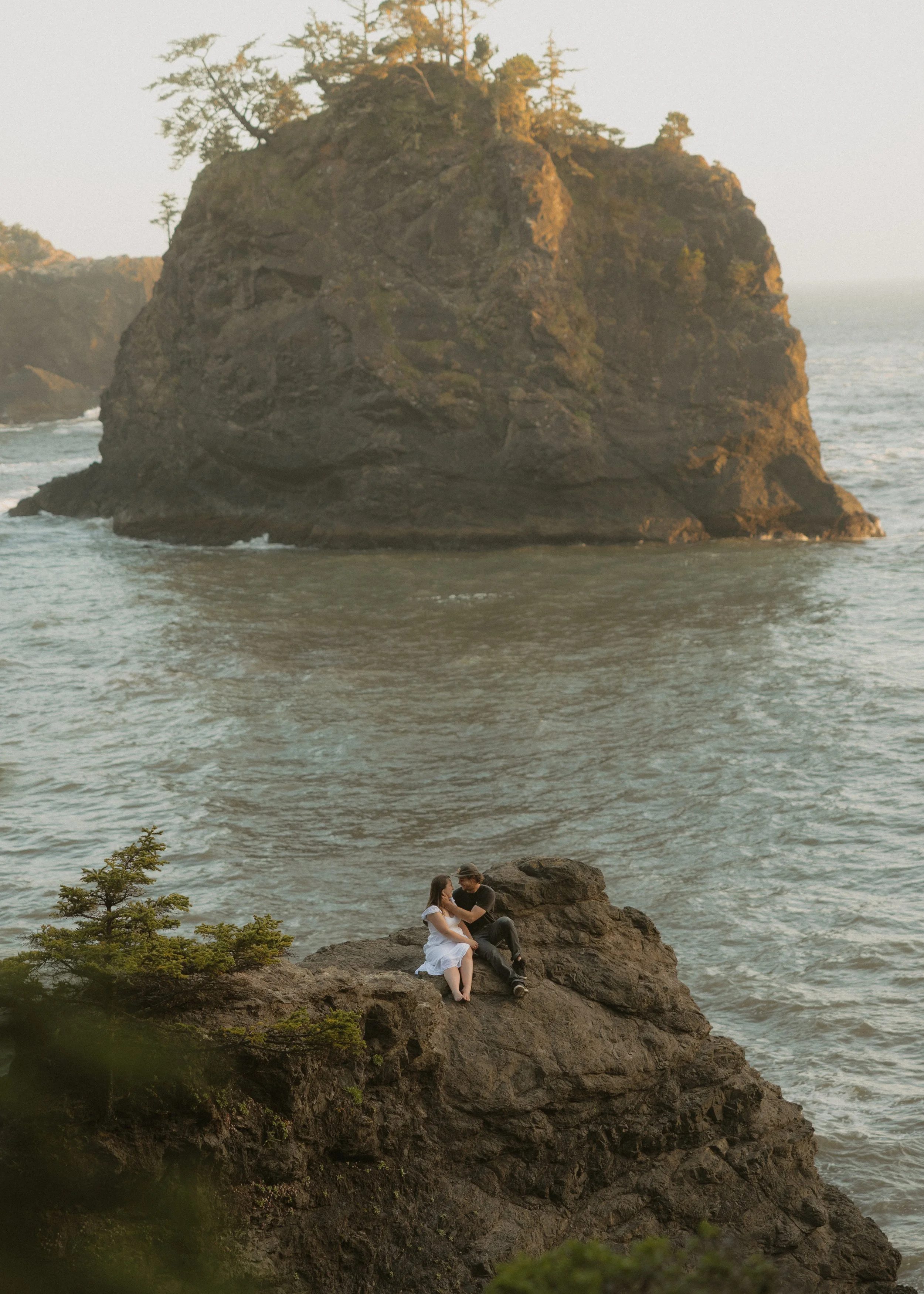 A couple sitting on rocks at the edge of the water, with a large rocky island in the background.