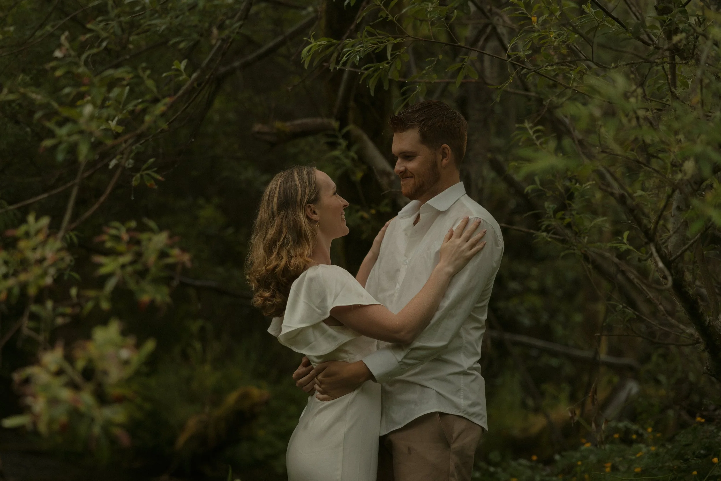 A couple is standing close together outdoors among tree branches, embracing and smiling at each other.