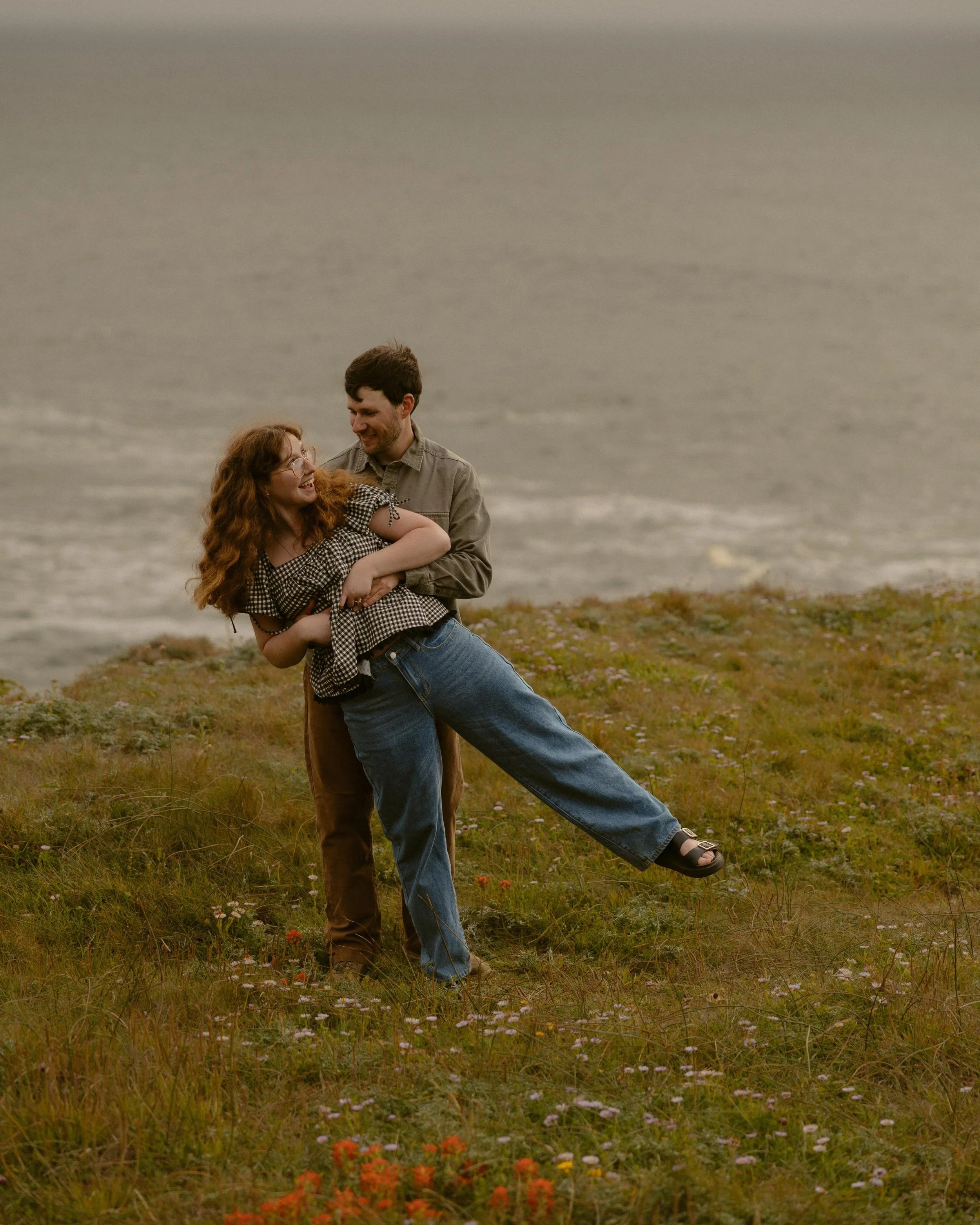 A couple stands in a grassy field near the ocean, with the woman being lifted off the ground by the man. They are smiling and looking at each other, with the woman wearing glasses, a checkered top, and sandals. The man is dressed in a beige jacket an