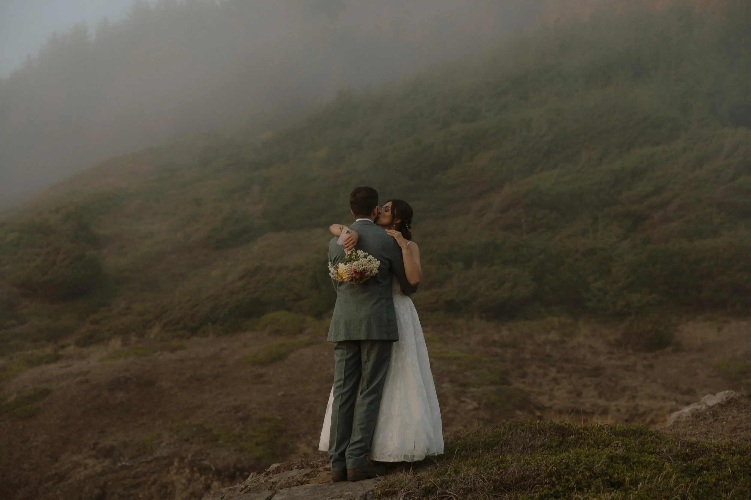 A bride and groom share a kiss outdoors on a foggy hillside. The groom holds a bouquet of flowers behind the bride, who is dressed in a white wedding gown. The scene is set in a natural landscape with green hills in the background.